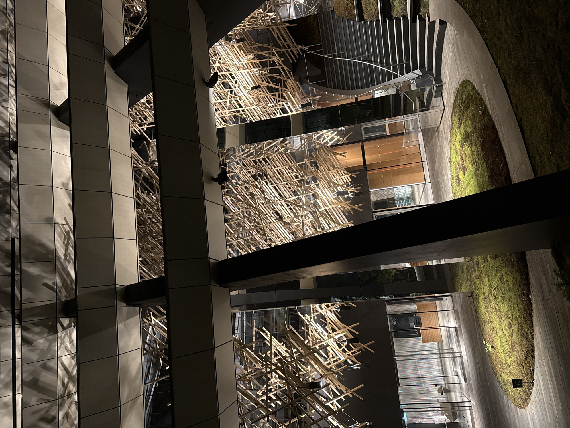 The image depicts an exterior view of a modern building complex in Vancouver, Canada, likely captured at night. The architectural style is characterized by clean lines, geometric forms, and the use of materials such as glass, metal, and possibly concrete. The building's facade features a combination of reflective and opaque surfaces, with vertical panels running along its height.
A prominent design element is the intricate wooden structure, resembling a latticework or scaffolding, suspended between sections of the building. The wooden elements are illuminated, casting light and shadow on the surrounding surfaces.
In the background, there is a landscaped area with a pathway and vegetation, suggesting a courtyard or outdoor space within the complex. Stairs are also visible, leading to different levels of the building. The overall environment appears to be well-maintained and aesthetically designed.
There are no people visible in the image. The focus is on the architectural details and the interplay of light and shadow in the urban environment. The lighting suggests that it is either dusk or nighttime. The building features a sign that reads "1556". A sign with a graphic on it can be seen next to a grassy area. The orientation of the image is rotated counterclockwise.