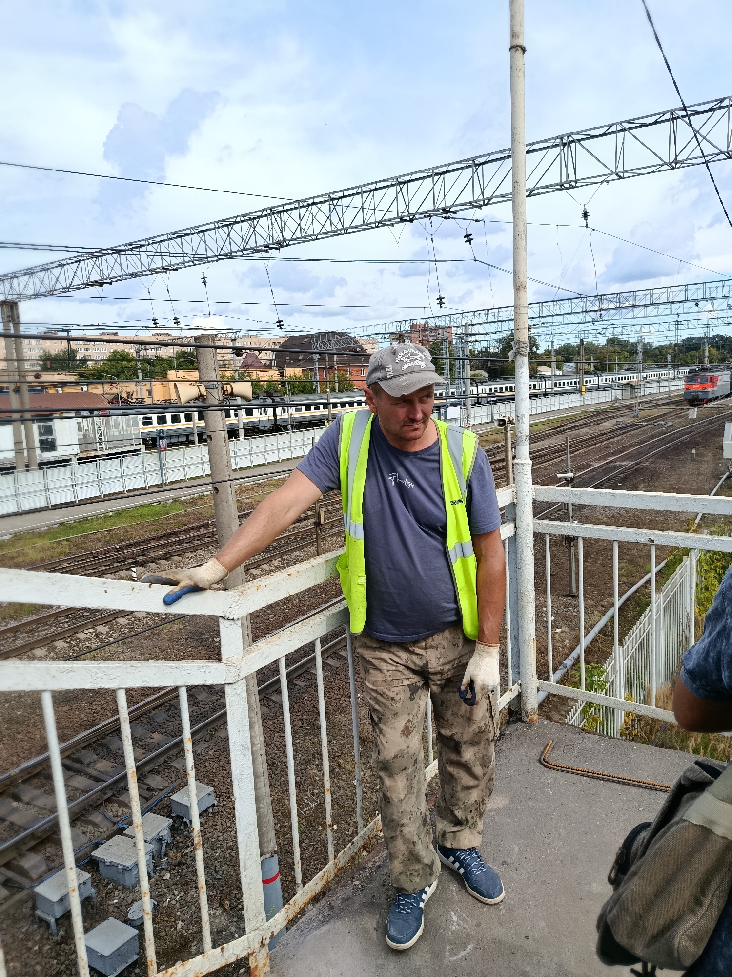 A man wearing a gray baseball cap, a dark blue t-shirt, a high-visibility yellow vest, camouflage pants, and blue sneakers stands on a pedestrian overpass, resting his left hand on the white metal railing. He is wearing white gloves. Behind him, train tracks and railway infrastructure are visible. Buildings and trees are in the background. The scene appears to be a railway station or junction in Orekhovo-Zuyevo, Russia.
