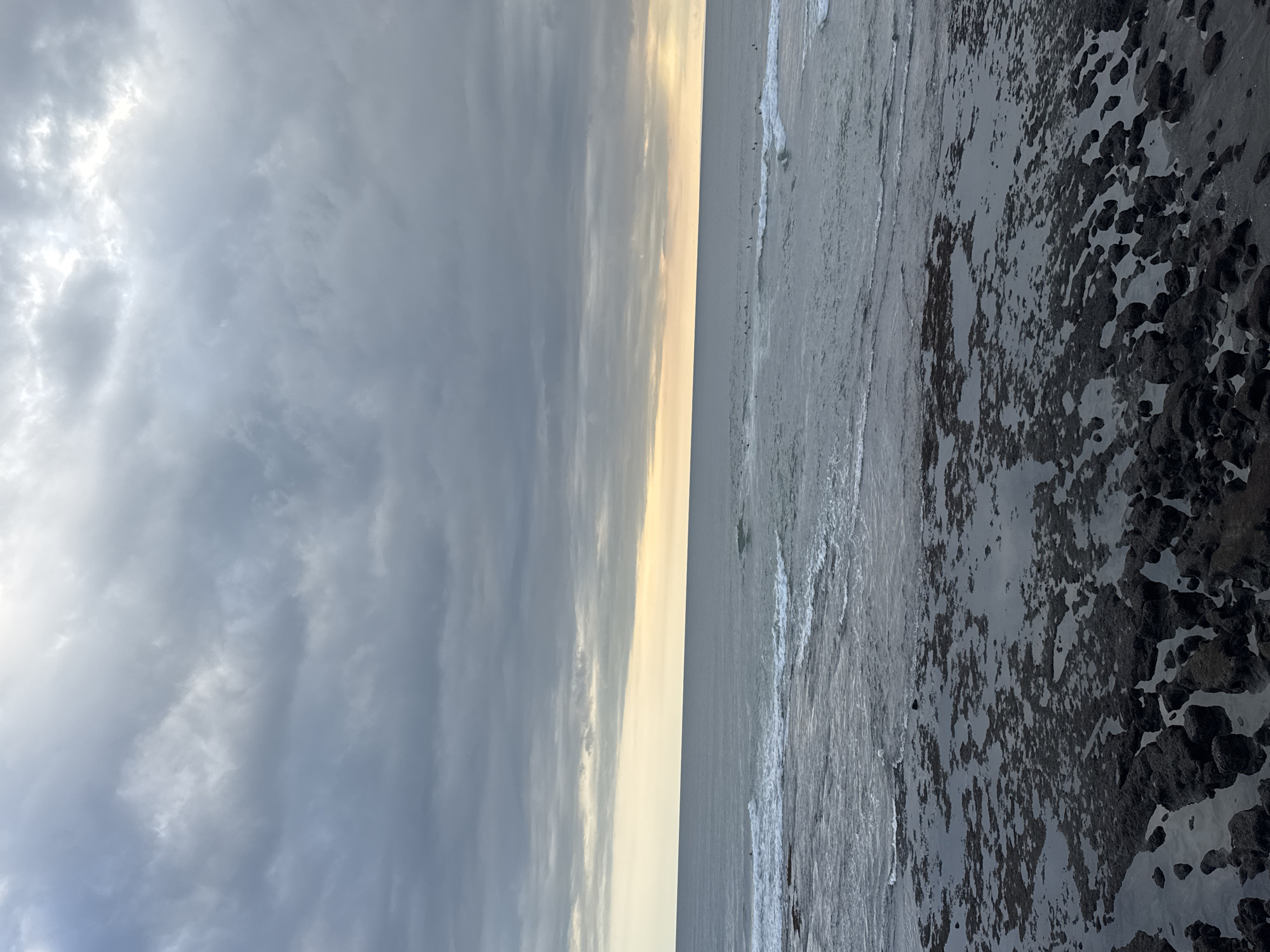 A landscape image of the beach in North Kuta, Indonesia. The sky is predominantly cloudy and the sun is setting on the horizon. The ocean is in the middle of the picture, with small waves near the shore. There are black rocks along the edge of the shore.