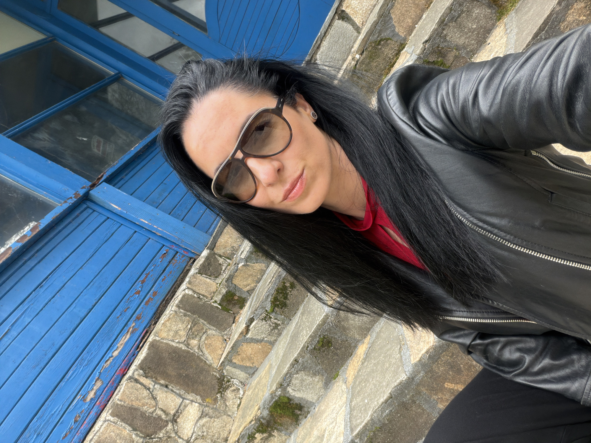 A woman is captured in a medium close-up, leaning or sitting outdoors, with the image taken from a slightly low, angled perspective. She is the central subject, positioned to the right of the frame, looking towards the camera. She has long, straight, dark hair that falls over her shoulders. She wears brown-tinted sunglasses with a distinctive light brown and gold frame, and a small stud earring is visible on her right earlobe. Her attire consists of a black leather jacket with prominent metallic zippers and a red shirt worn underneath.
Behind and to the left of her is a weathered blue wooden structure, likely an old window frame or set of shutters, showing significant peeling paint and exposed reddish layers beneath. To her right and extending into the background are stone steps or pavement, constructed from irregularly shaped, light-colored stones, some of which are covered with patches of green moss. The bright lighting suggests it is daytime, possibly sunny. No visible text is present in the image. The setting appears to be an outdoor area, possibly near an old building or structure in Pleven, Bulgaria, as indicated by the location context.
