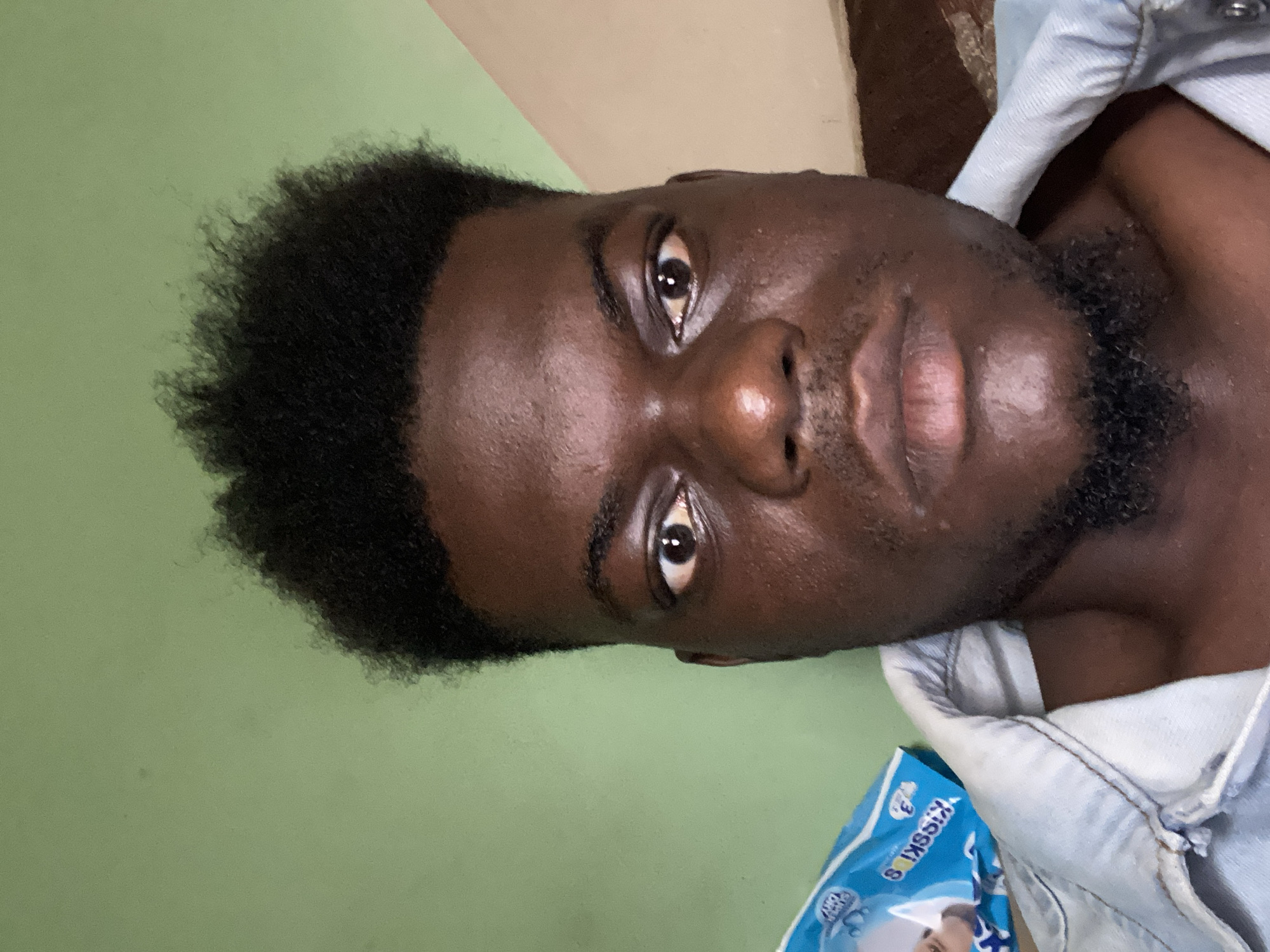 A close-up, slightly angled shot depicts a Black male with short, curly black hair and a short beard. His skin tone is a deep brown and he has dark eyes. He appears to be wearing a light blue denim jacket or shirt. Part of a blue and white package, possibly containing baby wipes or diapers, is visible near his shoulder. The background is a light green wall with a darker, beige area near the top. The image lacks discernible details to infer a location within a city.