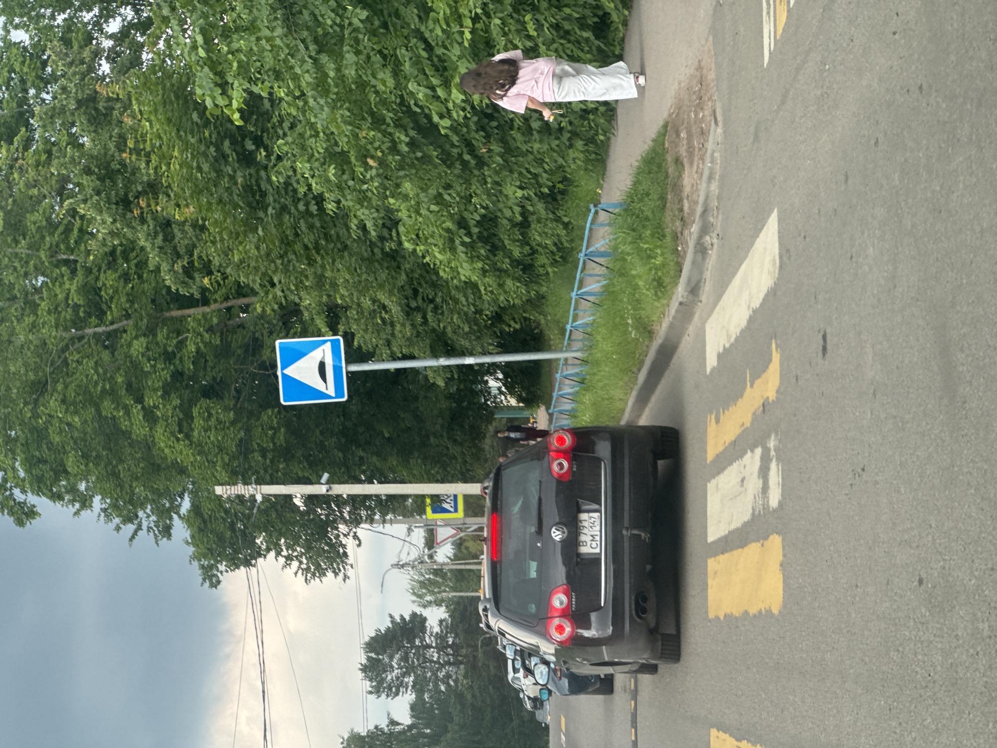 In Kuzmolovskiy, Russia, a medium-shot vertically oriented photo captures a street scene with pedestrians, vehicles, and natural elements under a cloudy sky, suggesting overcast weather.
The primary subjects are two people and several cars. A young woman with long, dark hair is seen walking on a sidewalk, wearing a pink t-shirt and light-colored pants. She appears to be holding a yellow object in her hand. Further down the street, a black Volkswagen Passat is parked, its brake lights illuminated. Behind it, other vehicles are visible, lined up on the road.
The setting is an urban street with a pedestrian crossing marked by yellow and white stripes on the asphalt. A blue and white traffic sign, indicating a pedestrian crossing, is prominently displayed on a pole. To the left of the road, a dense line of green trees provides a natural backdrop. A low blue fence runs alongside the sidewalk, bordering a grassy area. Overhead power lines are also visible.
The image appears to be taken during the daytime, but the overcast sky suggests it might be late afternoon or early morning, or simply a cloudy day. There are no strong shadows. The overall mood is neutral, depicting an everyday street scene.
Visible text includes the license plate of the Volkswagen, which reads "B 791 CM 147". The traffic sign is a standard international symbol for a pedestrian crossing.