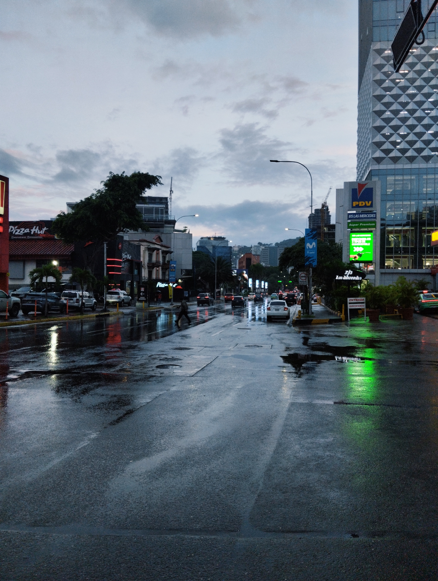 This is a street-level, eye-level, vertical, medium shot of a wet city street in Caracas, Venezuela, during overcast conditions, likely dusk or early morning. The wet asphalt reflects the surrounding lights and buildings.
On the left side of the street, a Pizza Hut restaurant with a red sign is visible. Several cars are parked or driving on the wet road. A lone pedestrian is walking across the street in the middle ground.
To the right, a tall modern building with a diamond-patterned facade dominates the background. Below this building, a PDV gas station sign is illuminated, along with green illuminated signage for "EIS LAS MERCEDES" and "ENTRADA BIENVENIDOS". Potted plants are positioned near the entrance.
The overall atmosphere is somber due to the weather and lighting conditions, with the wet pavement creating a reflective, somewhat abstract surface in the foreground and middle ground. The sky is cloudy and muted in color.