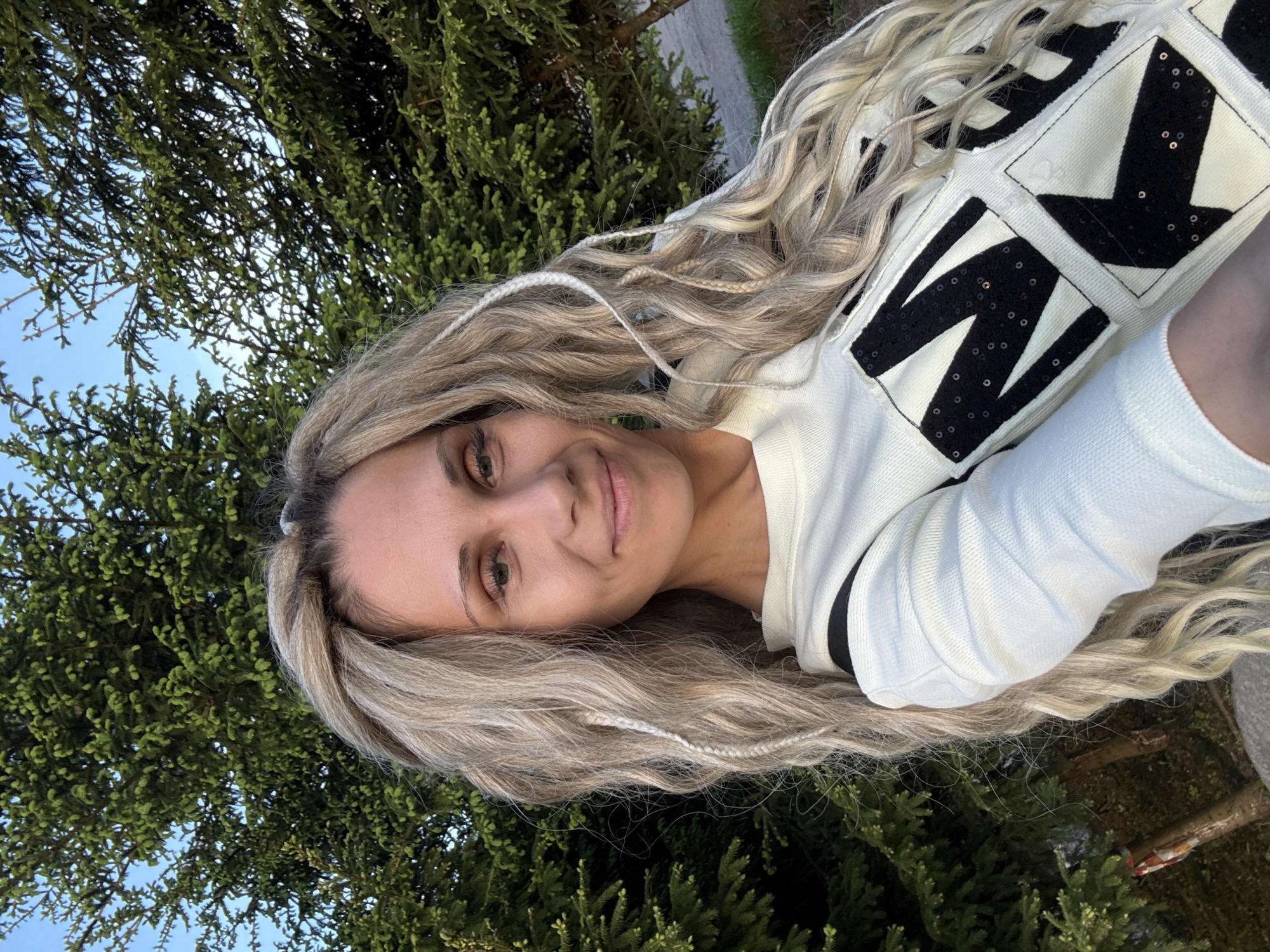 A woman with long, blonde, braided hair is posing for a selfie outdoors. She is wearing a light-colored sweater with black, sequined designs. The background consists of green foliage, possibly trees, with glimpses of blue sky visible through the branches. The location is possibly Rusanovo, Russia, but there is no identifiable feature in the image to confirm this.