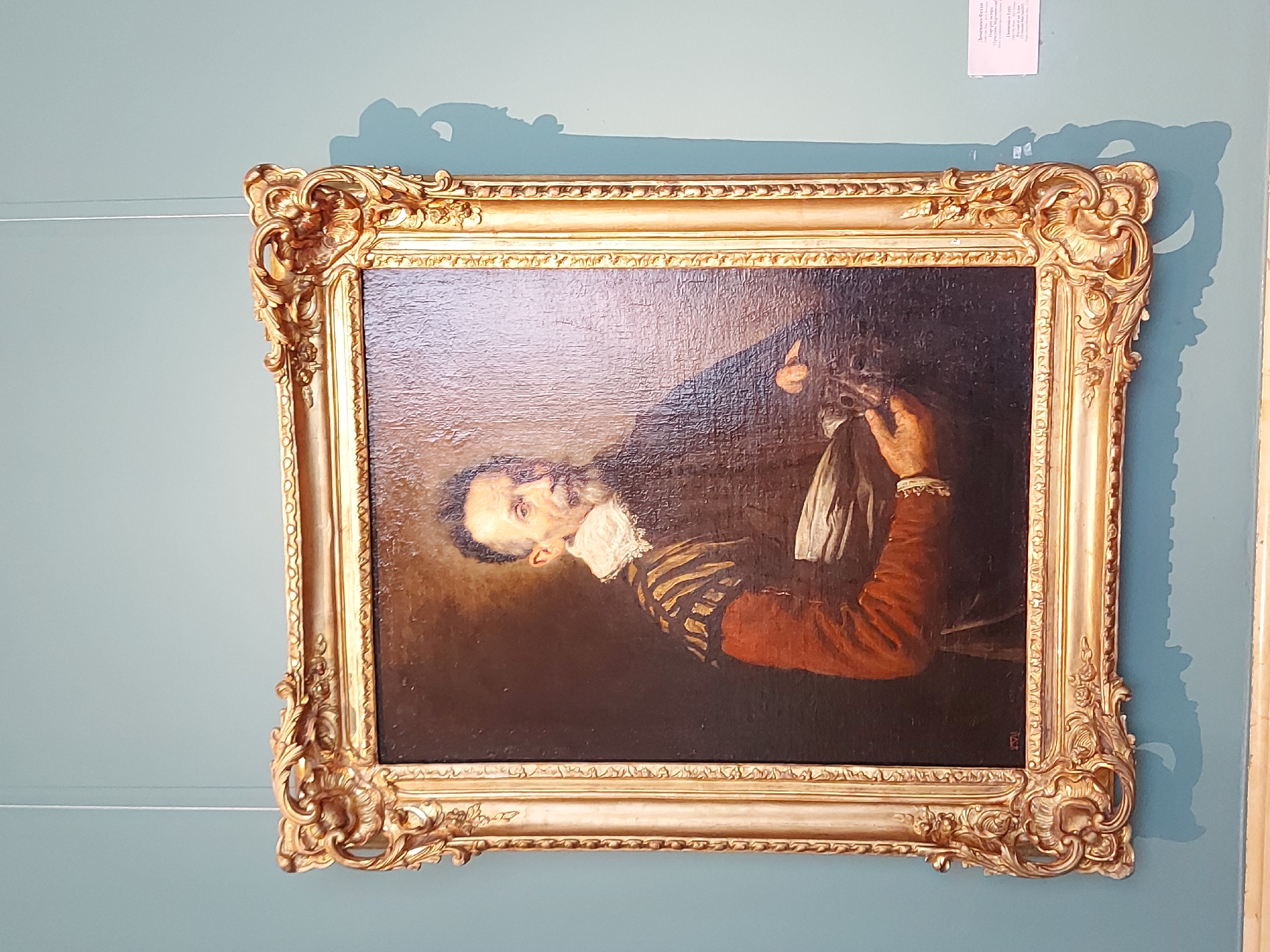 A framed painting hangs on a light blue wall. The painting is of a man with a beard and white collar, wearing a dark shirt with gold stripes and a red jacket. He is holding a white cloth. The frame is ornate and gold in color. A piece of paper is adhered to the wall above the top right corner of the painting. Thin wires are attached to the wall near the top left corner of the painting. The location is Unknown, Unknown.