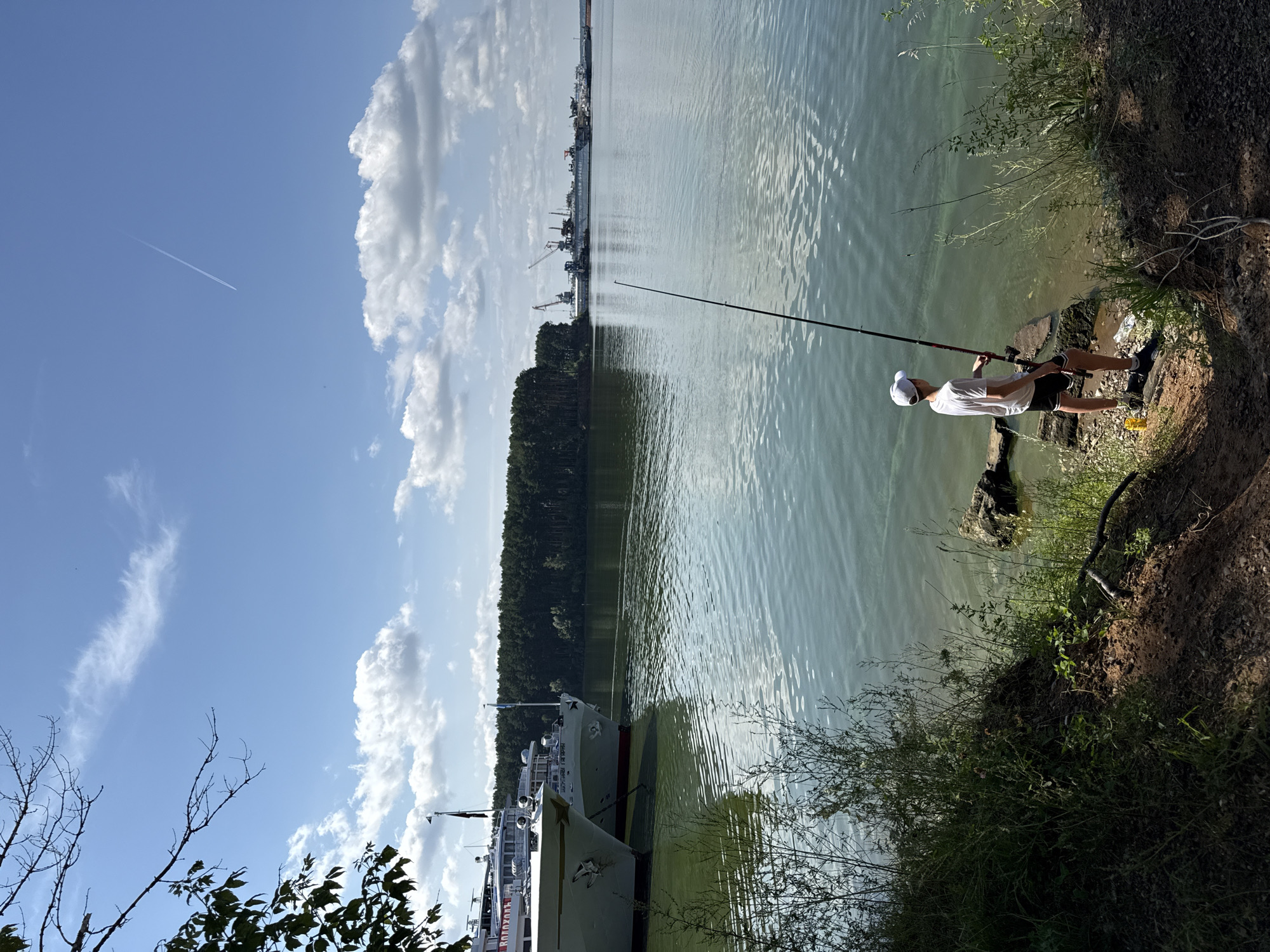A lone male, wearing a white t-shirt, black shorts, a white cap, and dark sneakers, stands on a rocky embankment of a wide, calm river, fishing. He is holding a fishing rod, with the line extended into the water. The river is bordered by a dense, dark green forest on the opposite bank. In the background, several large white passenger ships are docked, and further in the distance, a shipyard with cranes and other vessels is visible. The sky is a bright blue with scattered white clouds, and a contrail from an airplane stretches across it. The foreground shows a steep, earthy bank with overgrown vegetation. The image appears to have been taken on a sunny day, with the sun casting shadows on the embankment. This scene is likely situated along the Kama River in or near Chaikovsky, Russia, given the presence of docked ships and shipbuilding infrastructure commonly found on this waterway.