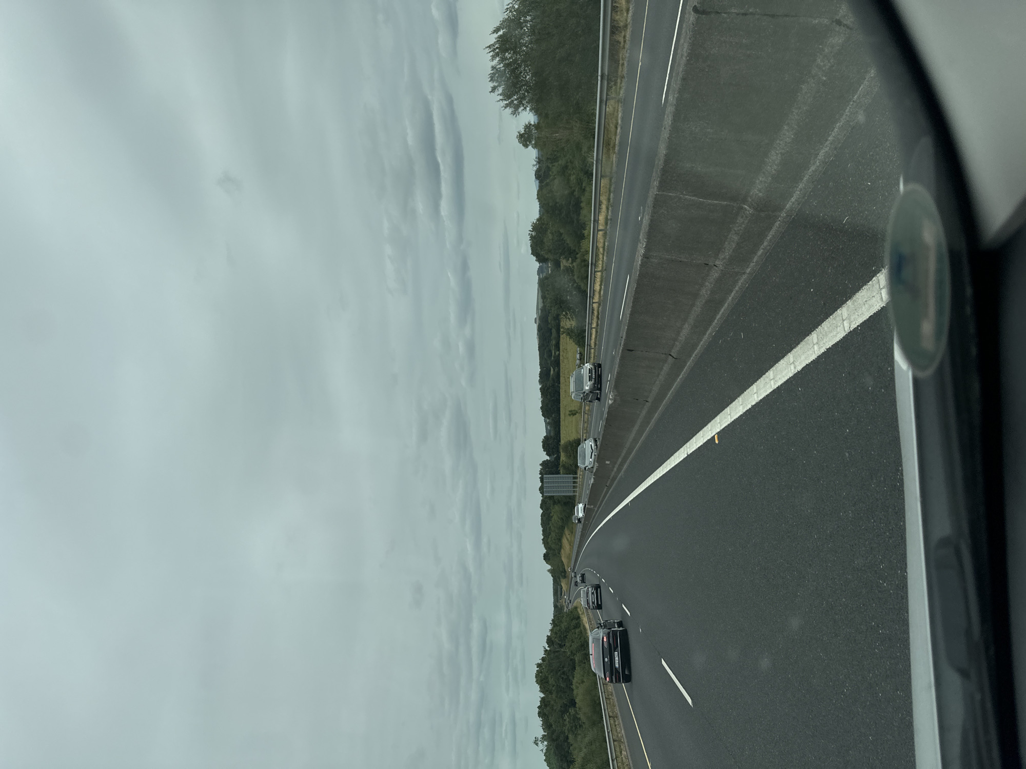 A wide-angle, eye-level exterior shot captures a view from a vehicle driving on a multi-lane highway under a cloudy sky. The highway, paved with dark asphalt, has clear white lane markings. Several cars are visible on the road, including a black sedan in the left lane and a white car further ahead. The highway is bordered on the right by a concrete barrier and a grassy verge, beyond which lies a landscape of rolling green hills and dense trees. The sky is overcast with a thick layer of gray clouds. The scene appears to be on the N24 highway, south of Stonehouse, Ireland.