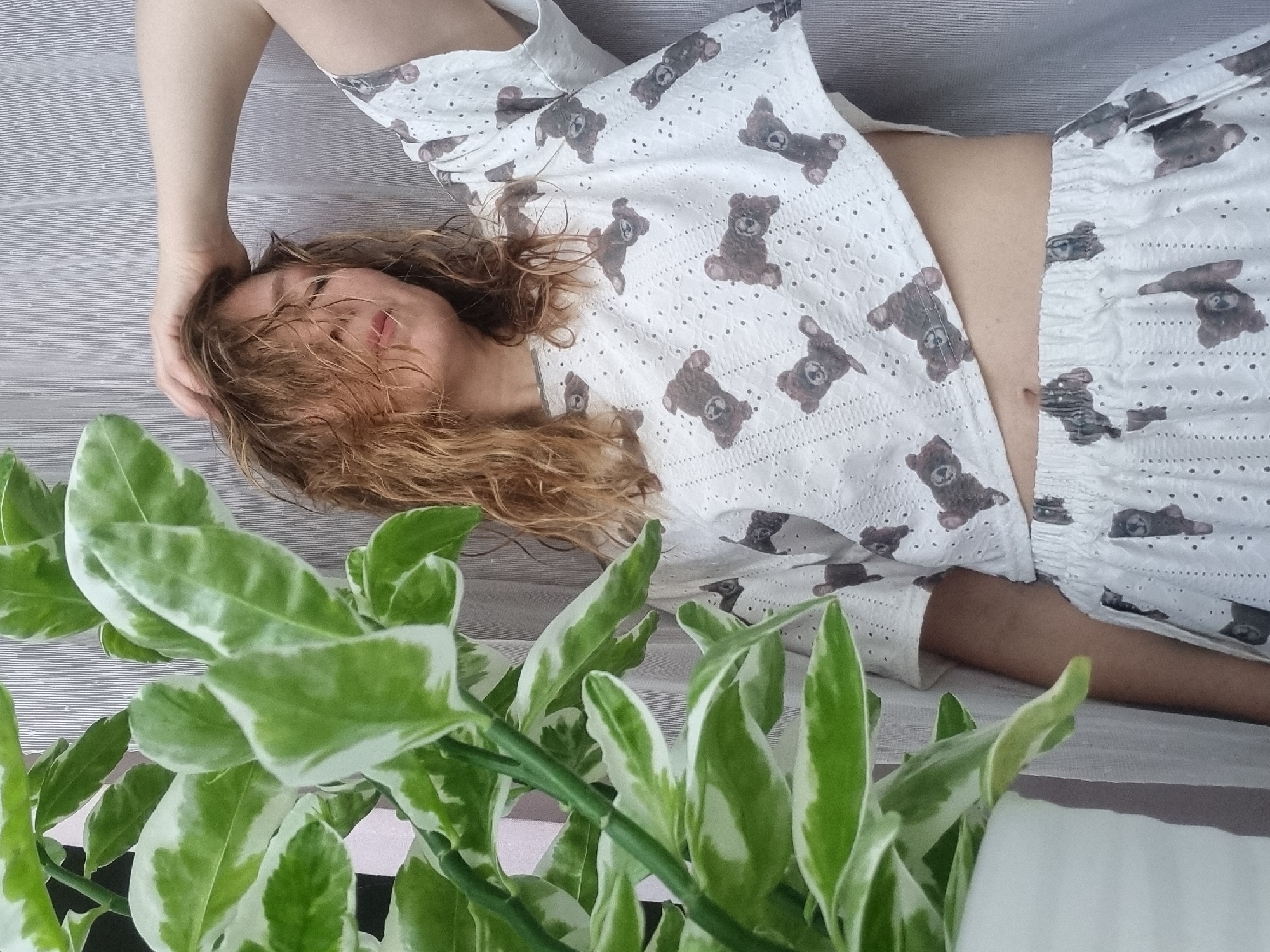 An overhead shot captures a person relaxing or resting, lying down on a light-colored surface with subtle horizontal stripes. The person's body is oriented diagonally across the frame, with their head positioned towards the top left. They have wavy, medium-length light brown or blonde hair that is somewhat disheveled and falls over part of their face. Their eyes appear closed or partially closed, and their lips are slightly parted, suggesting a state of rest or contemplation. One arm is bent, with the hand gently resting near or on the person's head, further indicating a relaxed posture.
The person is dressed in a two-piece white or off-white outfit, consisting of a short-sleeved crop top and matching shorts or lounge pants. The fabric of the clothing features a distinctive pattern of small brown teddy bears scattered across it, as well as an eyelet or embroidered texture with small perforations. The crop top exposes a section of the person's midriff, revealing their bare skin. The bottom garment also has the same teddy bear pattern and appears to have an elasticated waistband, visible where it meets the crop top.
In the foreground, partially obscuring the lower part of the person and the striped surface, is a vibrant green plant with variegated leaves. The leaves are distinctively shaped, with prominent bright green centers and lighter, almost white, margins. The plant's stems are also green, and it adds a natural, lively element to the scene. The lighting appears soft and diffused, possibly natural daylight, contributing to a calm and serene atmosphere. No text is visible in the image.