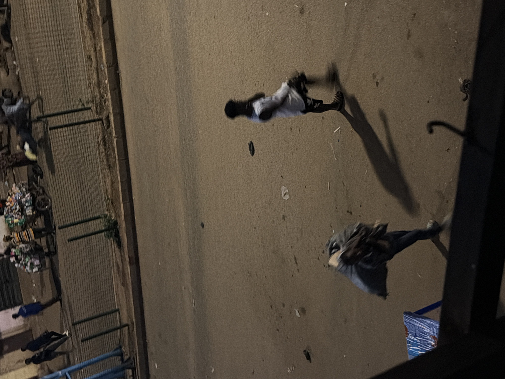 The image shows an overhead view of a street scene at dusk or night in Bukuru, Nigeria. The primary subjects are several people walking or standing along a paved road. On the left side of the image, a person in a blue shirt is walking, and further down, two people in darker clothing are also walking. Near the top left, a person in a light-colored shirt and yellow shoes is interacting with a cart laden with goods, possibly produce or merchandise.
In the middle of the street, a person wearing a white tank top and dark shorts is walking away from the camera. Further down the street, another individual is walking, wearing a dark jacket or hoodie and jeans. Their posture suggests they are moving at a brisk pace. Shadows are cast on the pavement, indicating an artificial light source or the fading daylight.
The setting appears to be an urban or semi-urban environment. The road is asphalt, and to the left, there is a raised area with a metal fence, behind which more people and what looks like a market stall or collection of goods are visible. There are also some discarded items and debris on the pavement. The overall lighting suggests it is either dawn or dusk, with artificial lights likely contributing to the illumination. No specific weather conditions are discernible, but the scene appears dry. No text is visible in the image.