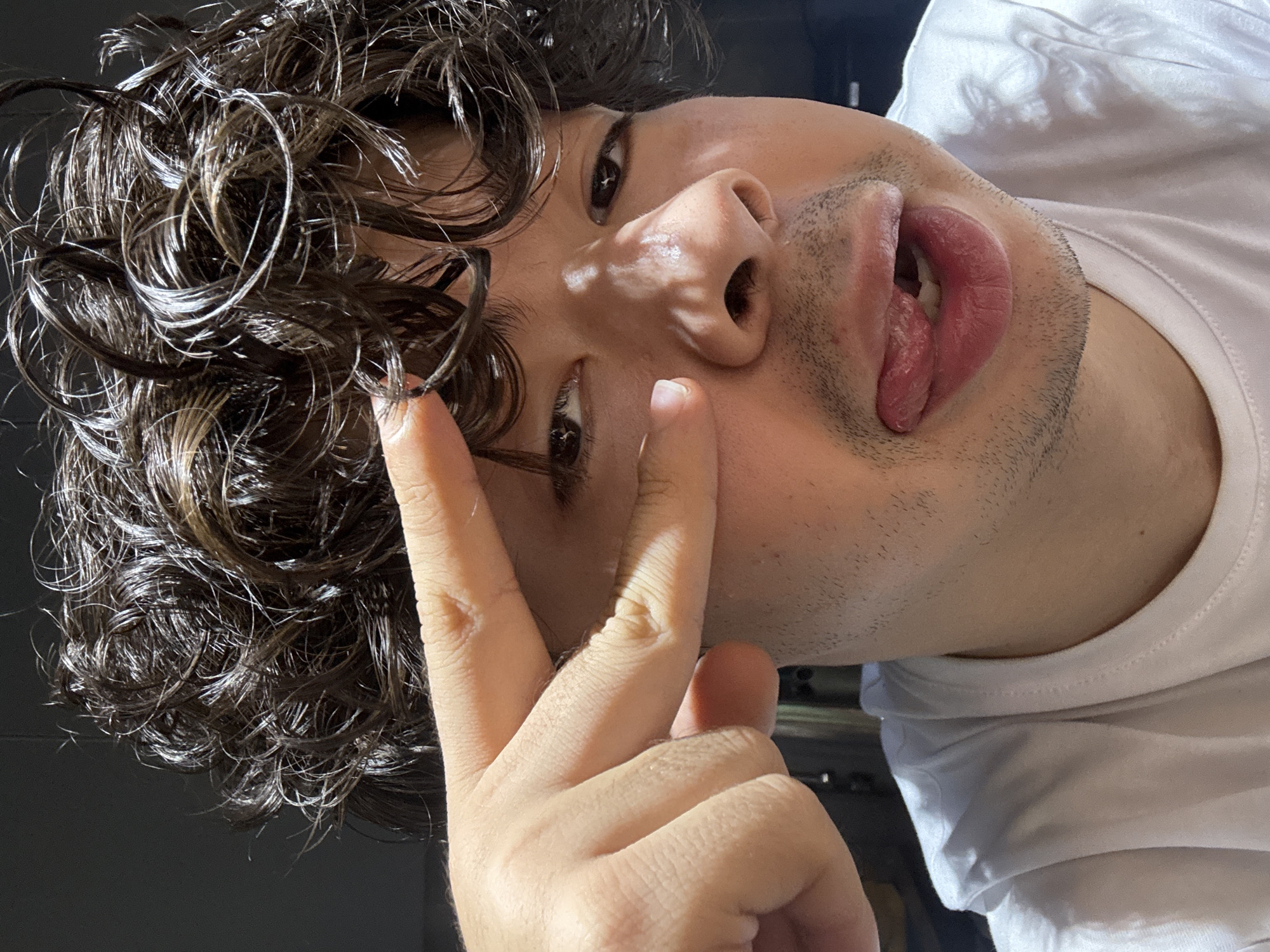 In a close-up, frontal shot, a person with curly brown hair, wearing a white t-shirt, extends their right hand, with the index and middle fingers touching their left eye. Sunlight casts shadows across their face, highlighting their nose and cheek. Their mouth is slightly open, with their tongue sticking out and touching their lower lip. The background is dark and indistinct, suggesting an indoor setting, possibly a home. The image does not contain any visual cues to determine its location in Tashkent, Uzbekistan.
