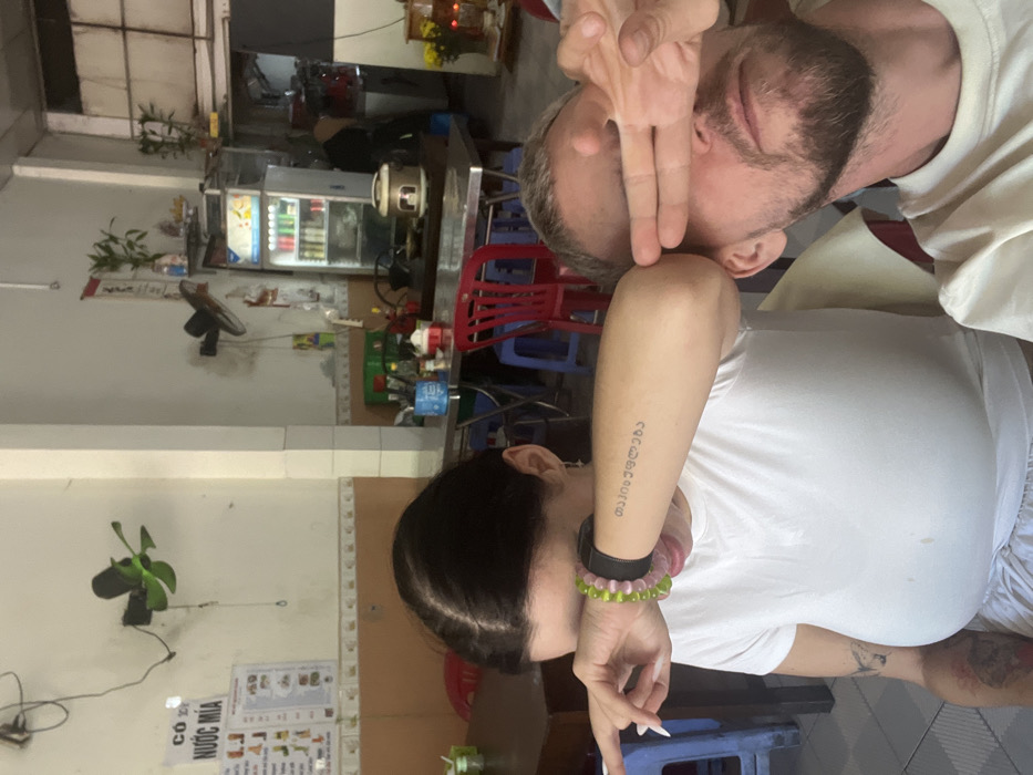 A man and woman are posing indoors, rotated sideways. The man, with a beard, holds two fingers up near his eye. The woman, with dark hair, has writing tattooed on her arm. They are in what appears to be a cafe. A refrigerator filled with drinks, a fan, and red plastic chairs are visible in the background. There's also a menu board on the wall.