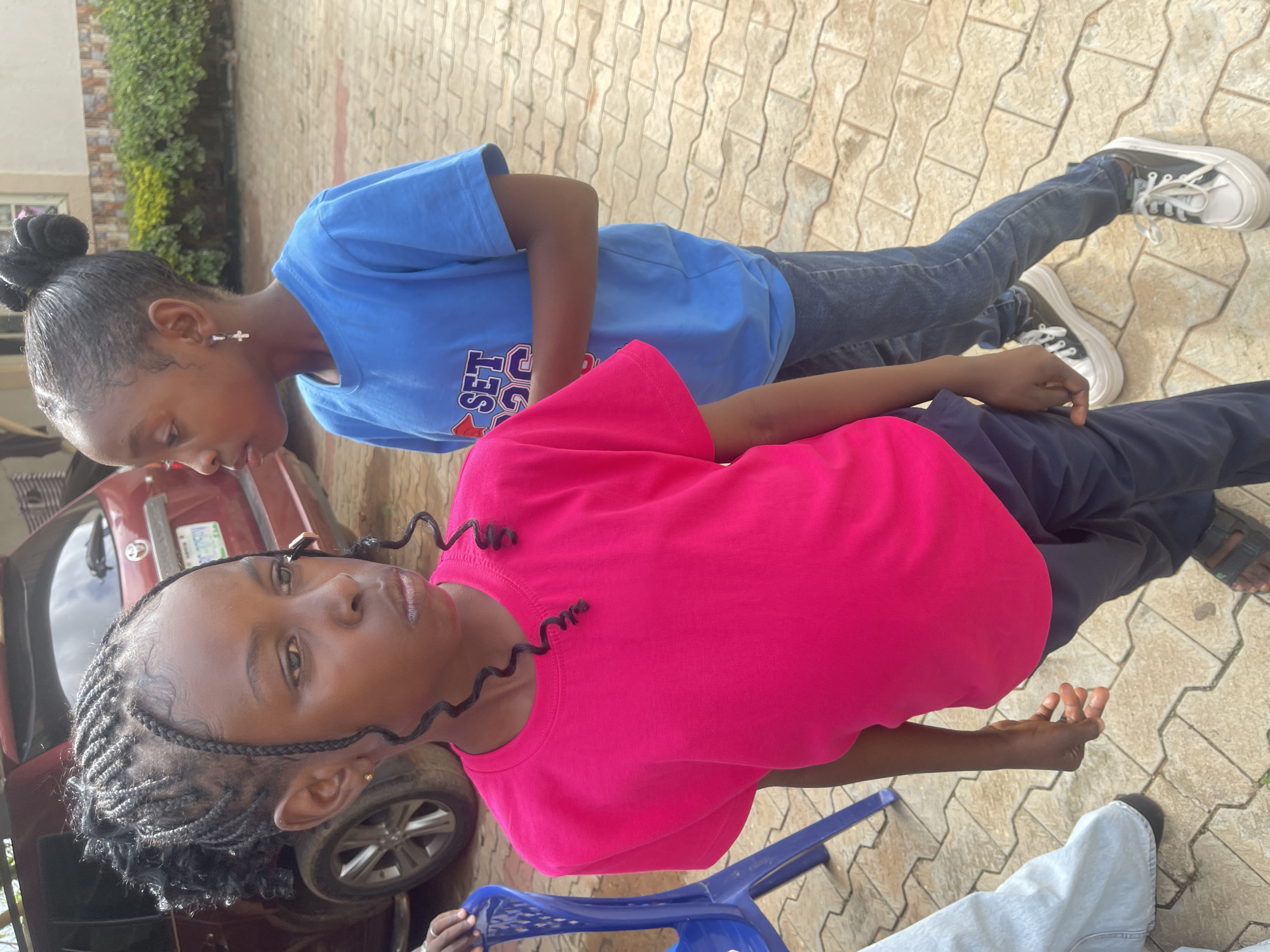 The image shows two young girls standing outdoors in what appears to be a residential or school compound in Gura Top, Nigeria. The girl in the foreground is wearing a bright pink t-shirt and dark blue pants. Her hair is styled in braids with curly tendrils falling around her face. She is looking to her left. Behind her, a second girl is wearing a blue t-shirt with a graphic print and dark denim jeans. She has her hair pulled up in a bun and is also looking to her left. Their stances suggest they are observing something outside the frame. In the background, a dark red SUV is parked, and a portion of a building with a patterned facade and green bushes is visible. The ground is paved with interlocking bricks. A blue plastic chair is partially visible on the left side of the image.