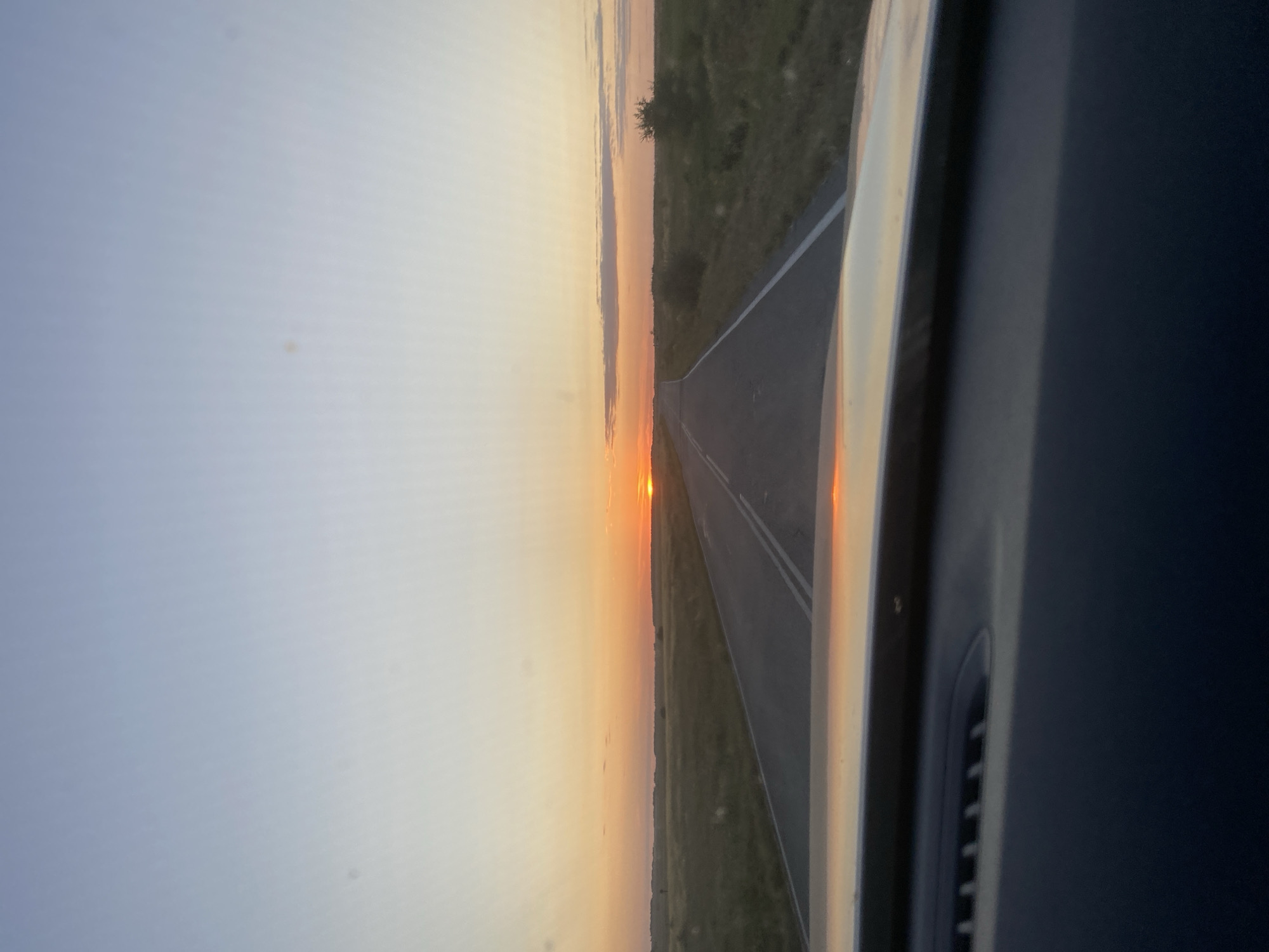 The image is taken from inside a vehicle looking out at a highway stretching into the distance. The sun is low on the horizon, casting a warm, orange glow across the sky and reflecting on the road. The landscape on either side of the road is a flat, grassy field with scattered bushes and trees. The sky is clear with a faint gradient of blue to orange. The front of the vehicle's dashboard is visible in the foreground, with the windshield reflecting the sunset. The location is identified as Абубакирово, Russia.