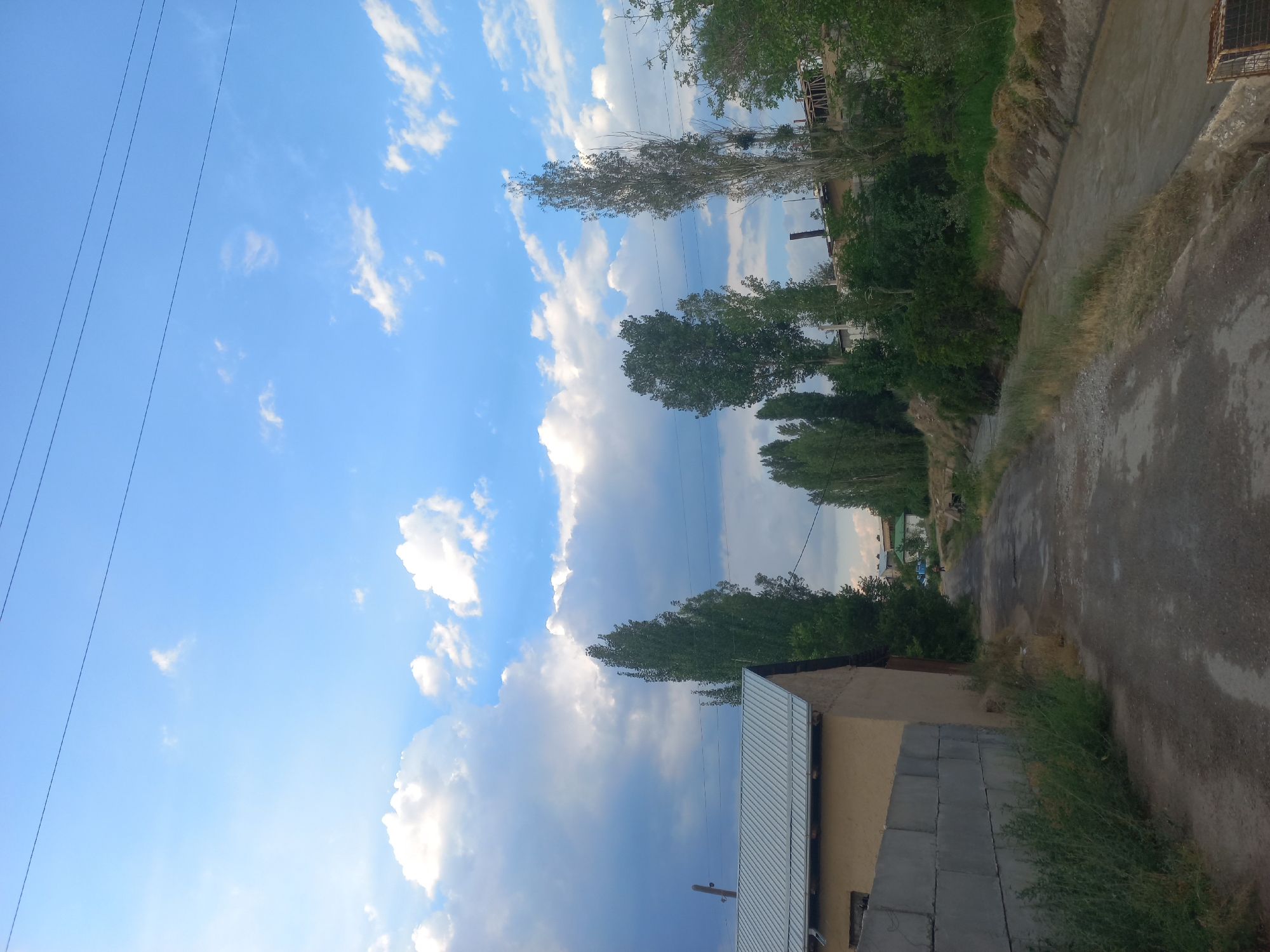 The image depicts an outdoor scene with a blue sky filled with scattered white clouds. In the foreground, there's a residential building with a beige exterior and a corrugated metal roof. Adjacent to the building, a wall constructed from grey concrete blocks is visible. To the right of the building, a dirt path or road is present, bordered by dry grass and vegetation. Further into the background, a row of trees lines the landscape, with denser foliage on the right side of the image. The sky suggests it is daytime, likely afternoon, given the lighting and cloud formation. The overall setting appears to be a rural or semi-rural area. There is no visible text in the image.