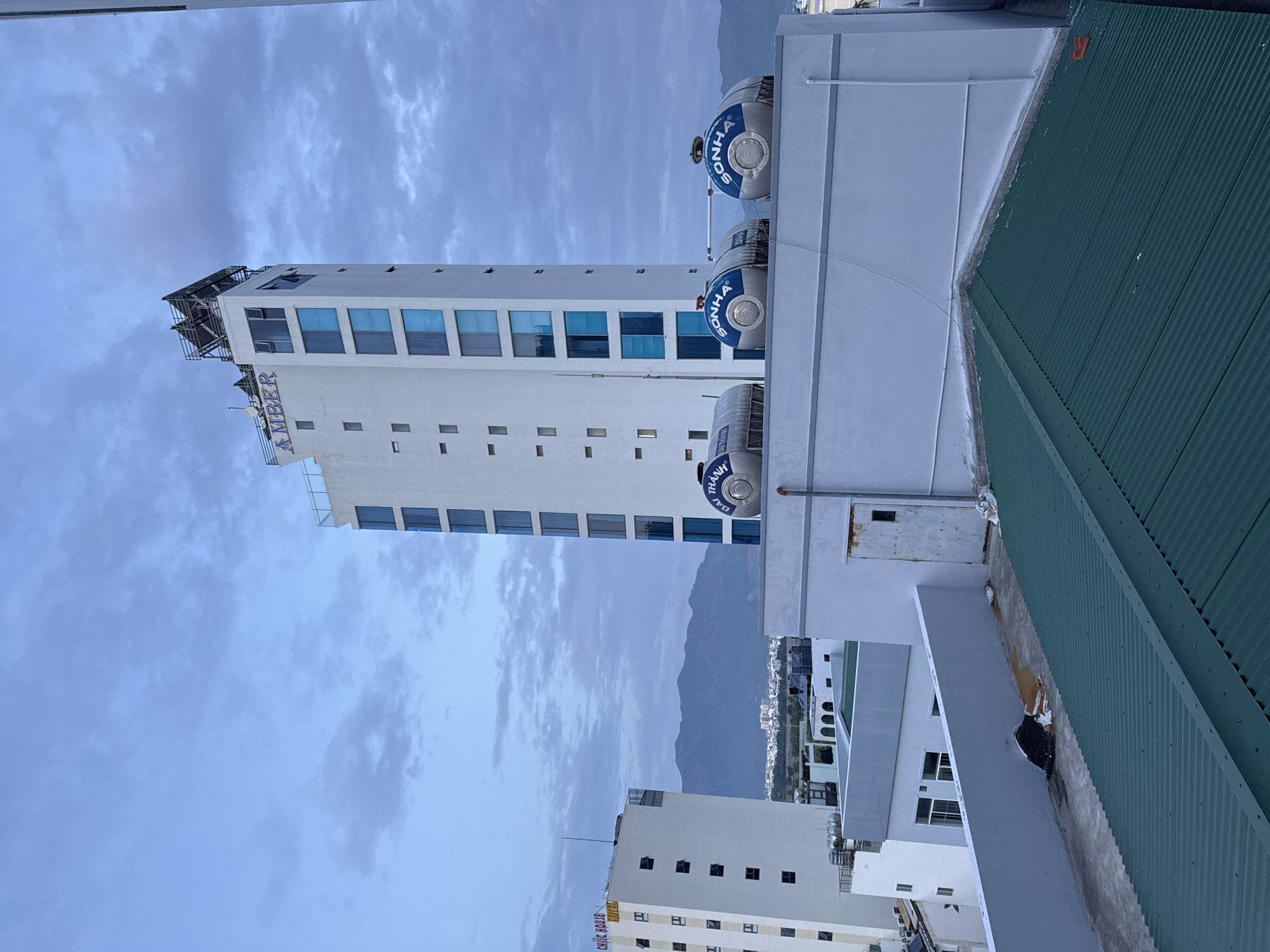 The image shows a view of the city of Nha Trang, Vietnam, with several buildings and mountains in the background. The sky is overcast with clouds. In the foreground, there is a green corrugated metal roof. Behind that, a white building with a gray wall and a rusty metal door is visible. Further back, a tall white building with blue-tinted windows and the word "AMBER" in blue letters on top stands prominently. Two large water tanks with "SONHA" written on them are on the roof of a lower white building. Another white building with "CHIC HOUSE HOTEL" written on it is visible to the left. Mountains and a hazy cityscape are in the distance.