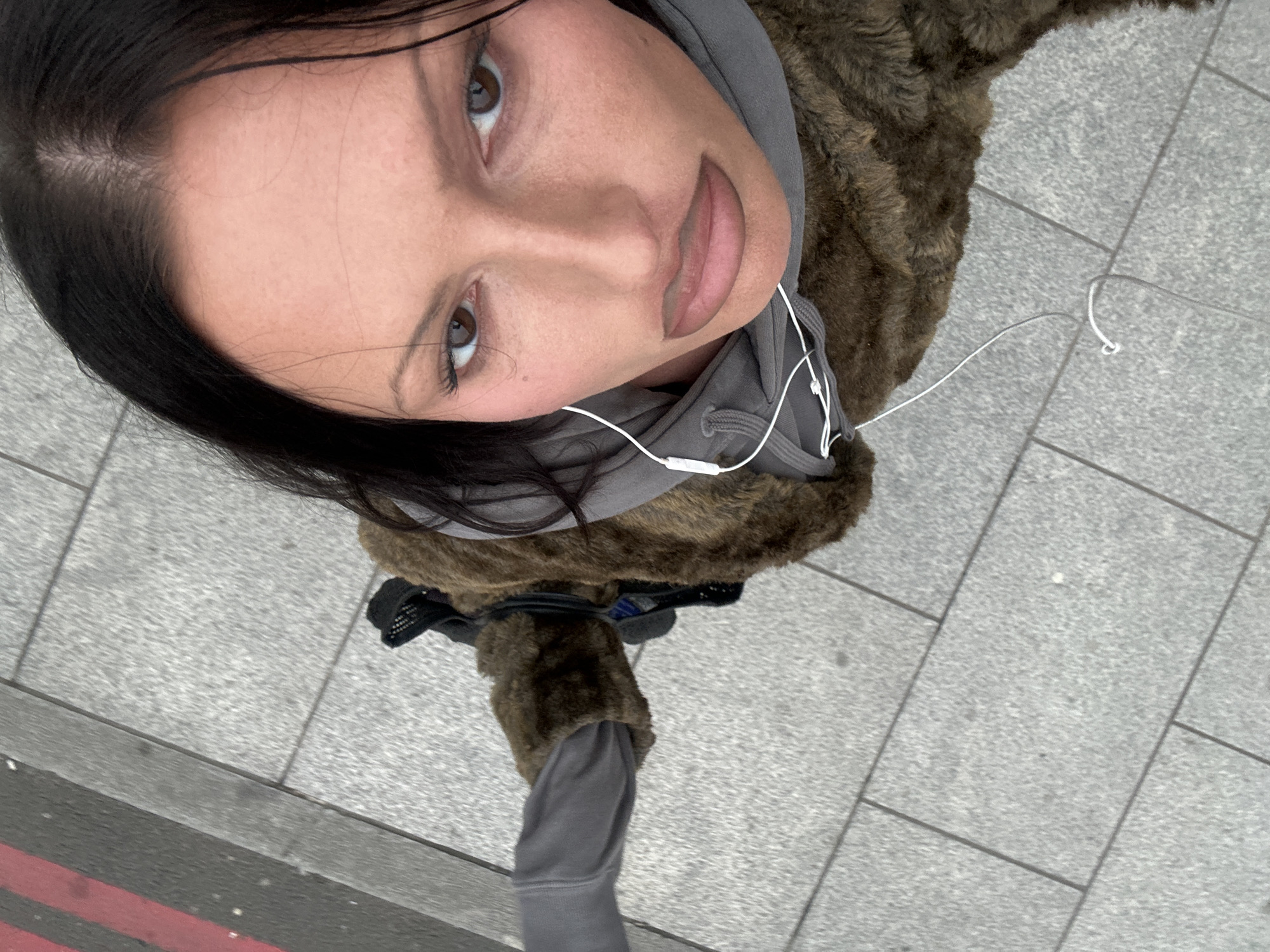 The image presents a high-angle, close-up view of an individual standing on a paved sidewalk in London, United Kingdom. The subject, appearing female, has dark hair parted centrally, brown eyes, and prominent facial features with visible makeup, including defined eyebrows and full, outlined lips. She wears a grey hooded sweatshirt underneath a textured, brown outer garment, likely a faux fur coat, with one grey sleeve visible. White wired earphones extend from her ears, draping across her chest. To the left of the light grey rectangular pavement slabs, a darker asphalt road surface is visible, distinguished by a red line bordered by a black line, indicating a "Red Route" characteristic of London's infrastructure. The scene is illuminated by ambient daylight.