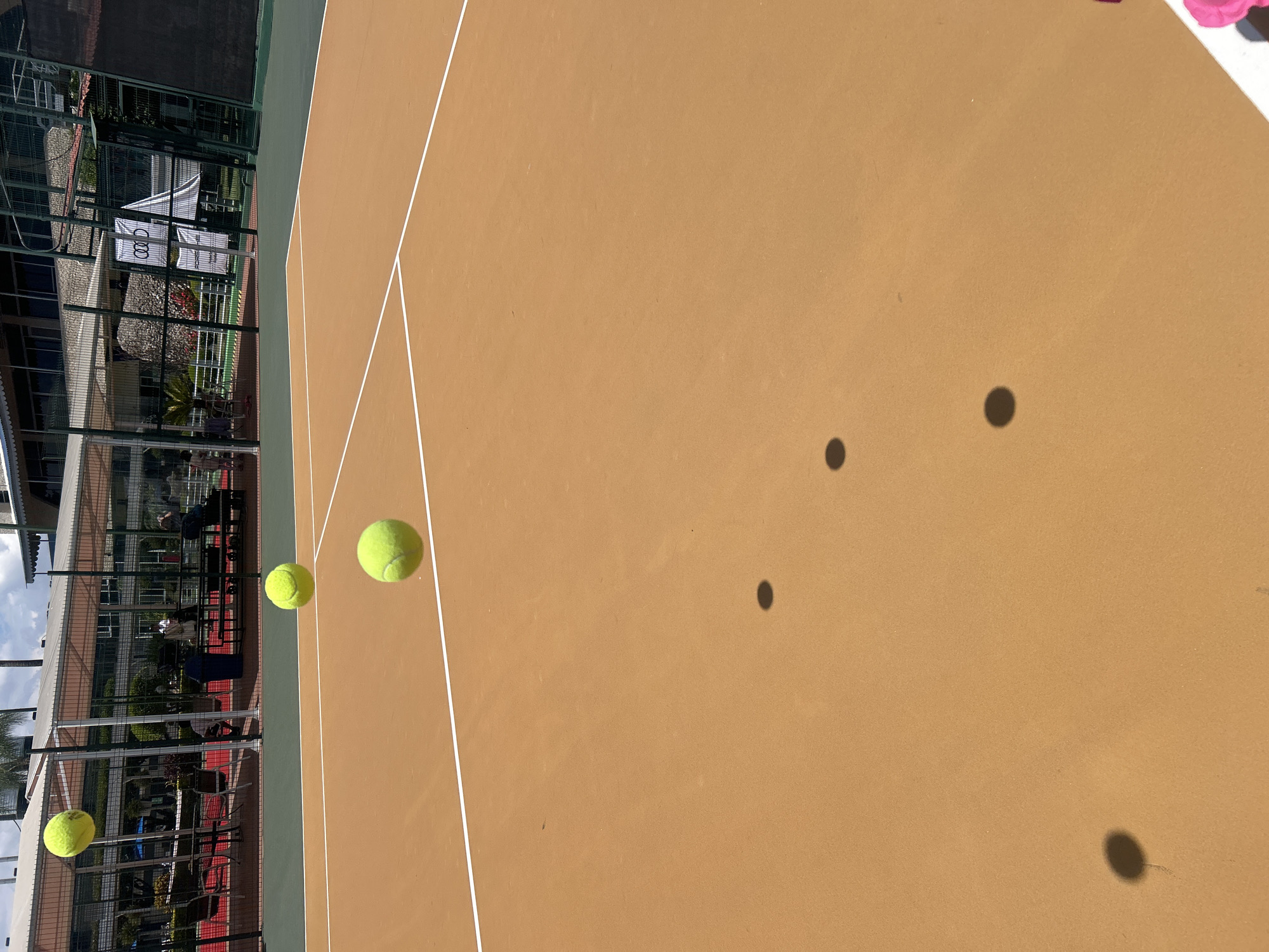 The image shows a tennis court with a tan playing surface and white lines. Three tennis balls are in the air, casting shadows on the court surface. In the background, there is a building behind a chain-link fence with an advertisement visible. Based on the advertisement, the location is likely the Drive Tennis Academy in Puebla, Mexico.