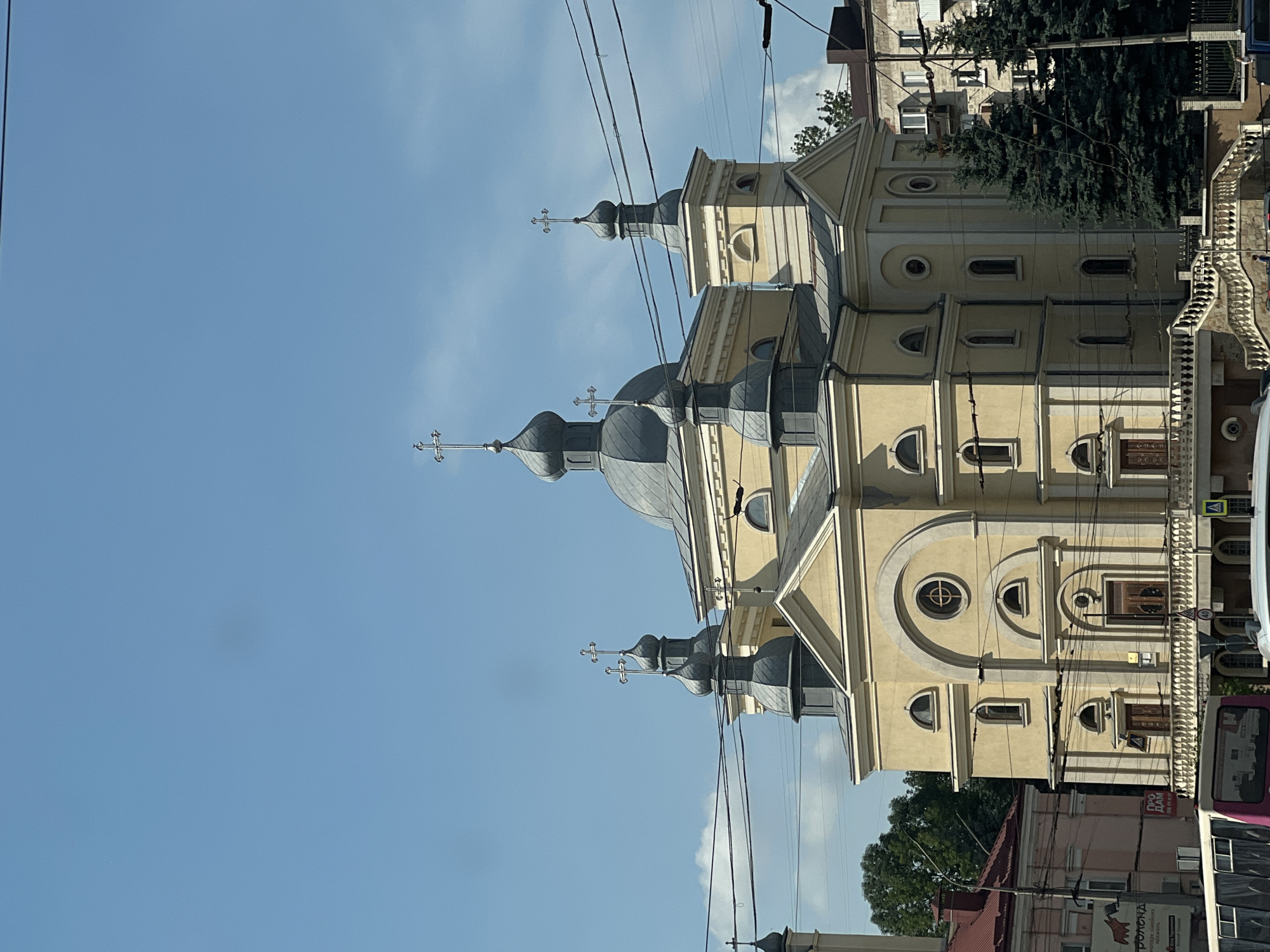 The image captures a view of the Ternopil Theological Seminary building in Ternopil, Ukraine. The prominent structure is a light yellow building with a domed central section and several smaller domes topped with crosses. The building exhibits classical architectural elements, including arched windows and decorative moldings. In the foreground, a bus is partially visible on the left, and a white car is parked on the right. Power lines crisscross the sky above the building. The sky is a bright blue with scattered white clouds. To the left of the seminary, a portion of a pink building with a red roof is visible, and to the right, a modern apartment building with balconies and windows can be seen. Several tall, dark green evergreen trees are positioned in front of the apartment building. The scene is bathed in sunlight, casting shadows on the building and the surrounding area.