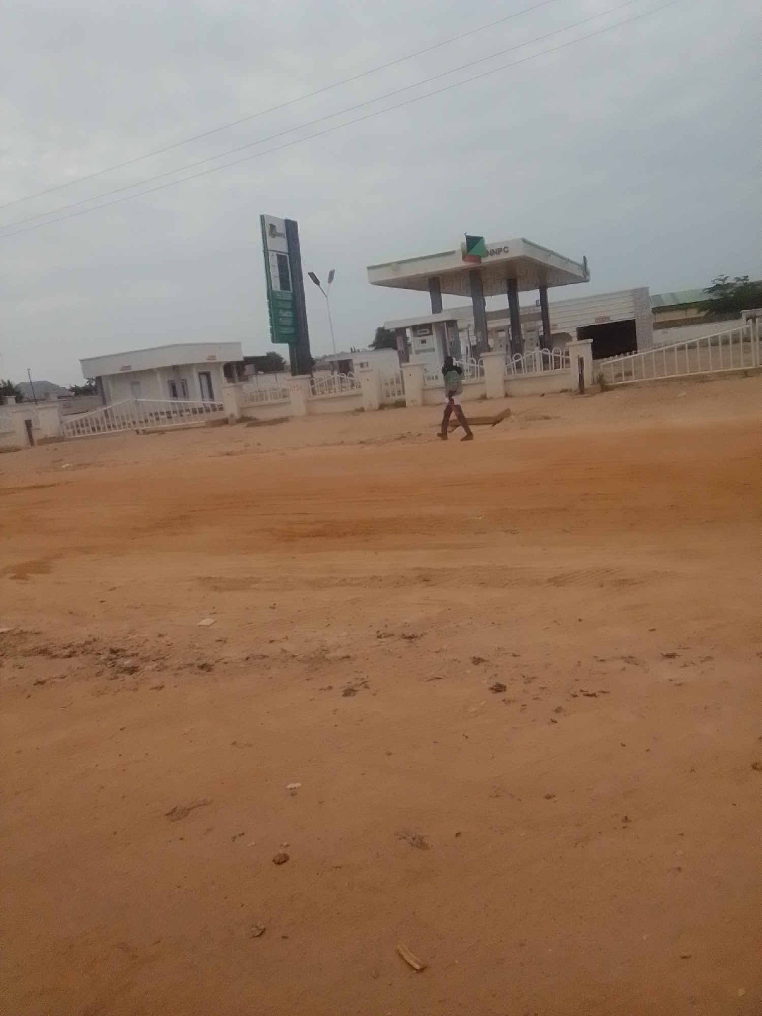 The image depicts an outdoor scene in the city of Gombe, Nigeria, featuring a NNPC gas station. In the foreground, a sandy, reddish-brown terrain is visible, with tire tracks suggesting vehicle movement. A person is walking across the terrain from left to right, carrying a broom or similar long, thin object. Behind the person, the NNPC gas station complex is situated. This includes a white canopy supported by pillars, indicating fuel pumps beneath. A tall, green and white NNPC signage stands to the left of the canopy. To the left of the gas station structure, a white building with multiple windows and a white fence surrounding the property can be observed. The sky is overcast, contributing to a subdued lighting condition. The overall impression is that of a functional, everyday scene at a Nigerian gas station.