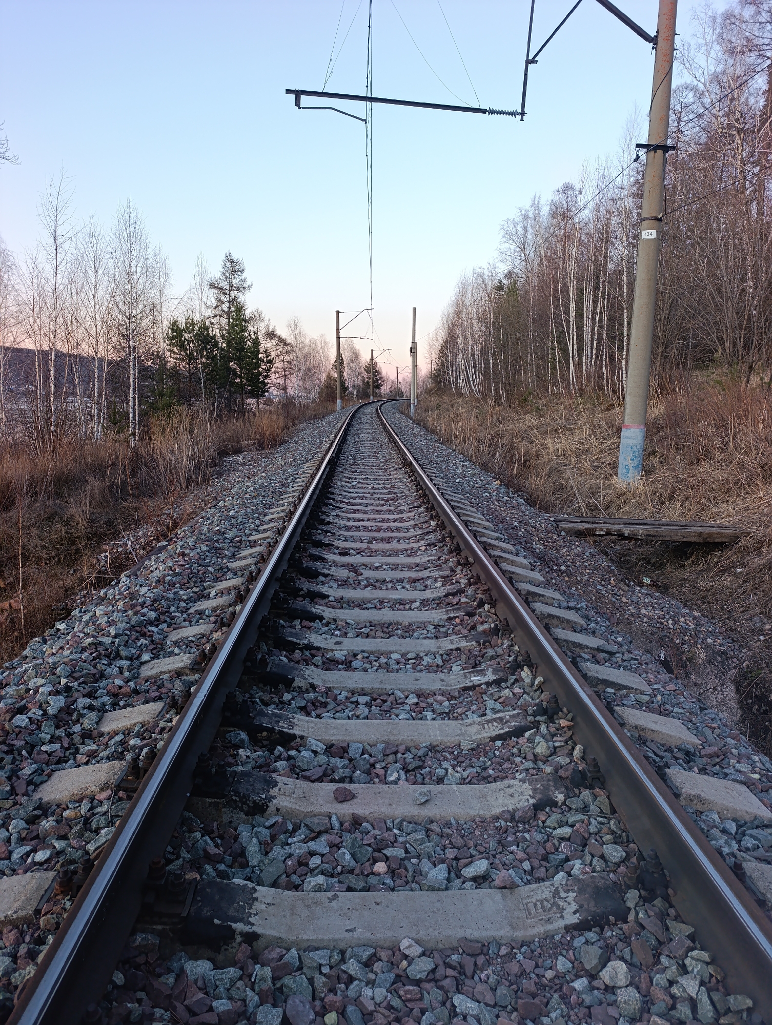 The image depicts a straight section of railway tracks running through a wooded area. The tracks are bordered by gravel and concrete sleepers. Overhead power lines and poles run parallel to the tracks. Trees line both sides of the tracks, with a mix of bare and sparsely leaved trees. The sky is visible in the distance, with a hint of sunrise or sunset. The lighting suggests it is either early morning or late afternoon.
Given the provided context and known locations, it is possible the image depicts tracks in or near Krasnoyarsk, Russia, along a railway line. However, without further information, the precise location cannot be definitively confirmed.
