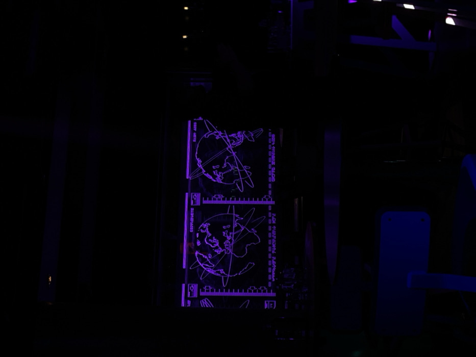 The image displays a dark, low-light setting, likely indoors, dominated by a vibrant purple glowing display panel. The panel features intricate line art, primarily consisting of two circular diagrams of globes or spheres, each encircled by multiple orbital rings and intersected by grid lines. Continents are discernible on the globes.
To the left of these diagrams, vertically oriented text is visible, including "GEOMAGNETIC" and possibly "GEOSPHERE". On the right side, further vertically oriented text reads "0327 METS", "SEN SOGUE GLOBE", and partially obscured "LOW RADIAL ARRAY". Below and between the globes are several horizontal scales or readouts, indicated by small glowing lines and numbers, resembling technical or scientific instrumentation displays.
The surrounding environment is largely shrouded in darkness, with only faint outlines of structures or equipment visible, tinted with dark blue and purple hues, suggesting additional lighting or reflections. The scene presents a high-tech or futuristic aesthetic, emphasizing the illuminated display against a dark backdrop. No people or overt activities are visible. The time of day appears to be night or an artificially lit dark space. The overall impression is one of data visualization or scientific monitoring.