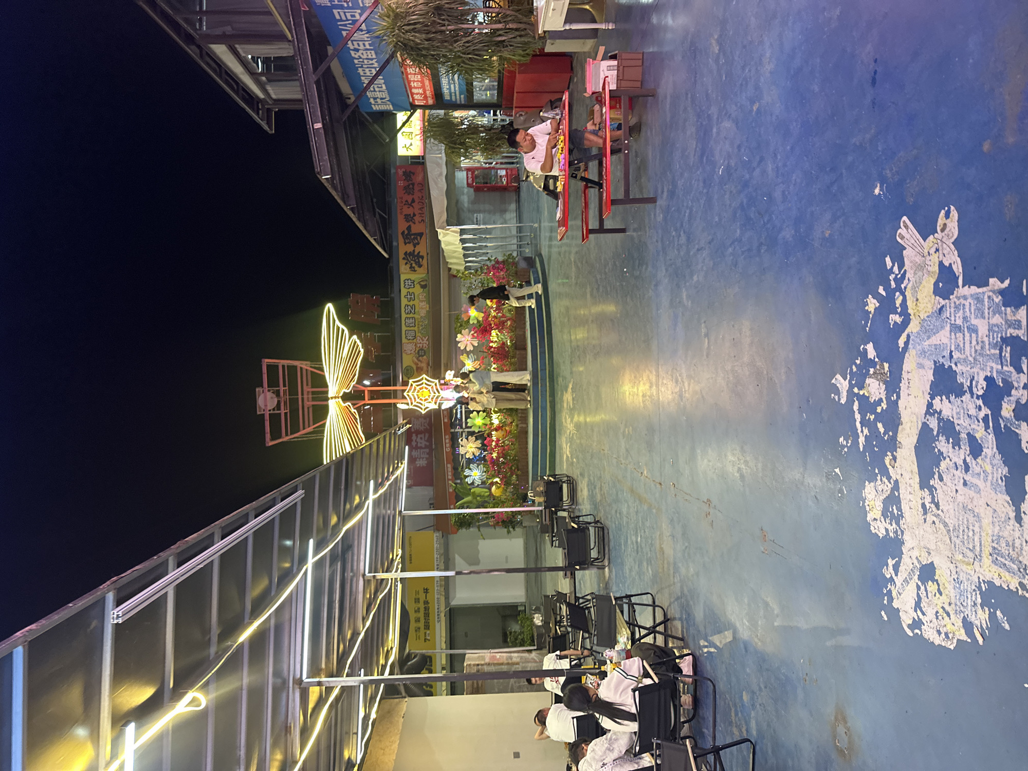 Here is a forensic description of the image:
**Setting:** The image shows an outdoor public space, seemingly in the evening or at night, in the city of Chongqing High-tech Zone, China.
**People:** Several people are visible in the scene, either seated or standing. One man is sitting at a red table with items on it. Other people are seated in black chairs. Some people are standing further back, looking at decorative displays.
**Objects:** The area is decorated with illuminated structures, including a butterfly and other illuminated displays. There are rows of black chairs set up. The surface appears to be a blue-painted area, possibly a basketball court. The background features storefronts with signage. There are metal structures overhead with attached lights.
**Actions and Interactions:** The people in the image seem to be relaxing and spending time outdoors. The man at the table appears to be engaged in an activity at the table. Some of the other people are seated in chairs, and others are looking at the decorations.
**Environment:** The environment is generally well-lit due to the overhead lights and illuminated displays. The area has a mix of natural and constructed features, with buildings in the background and decorative elements in the foreground.