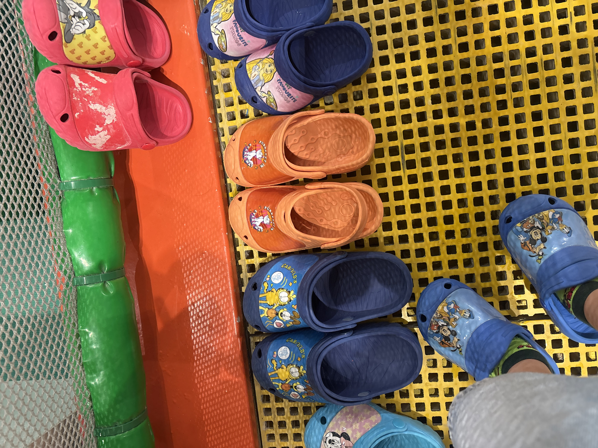 The image shows a close-up, top-down view of several pairs of children's Crocs shoes lined up on a yellow, grid-patterned surface. The shoes are diverse in color and design, featuring popular cartoon characters like Garfield, Mickey Mouse, and a character that appears to be from a Tom and Jerry cartoon. Some pairs are pink, others are orange, blue, and a light cyan. Two pairs of blue Crocs with Mickey Mouse characters are being worn by a person, indicated by their visible feet and patterned socks. In the background, a green, inflatable tube is visible, partially obscured by a red platform. The overall environment suggests an indoor play area or a similar recreational facility. The specific location in Dubai, United Arab Emirates, cannot be determined from the visual information provided in the image.