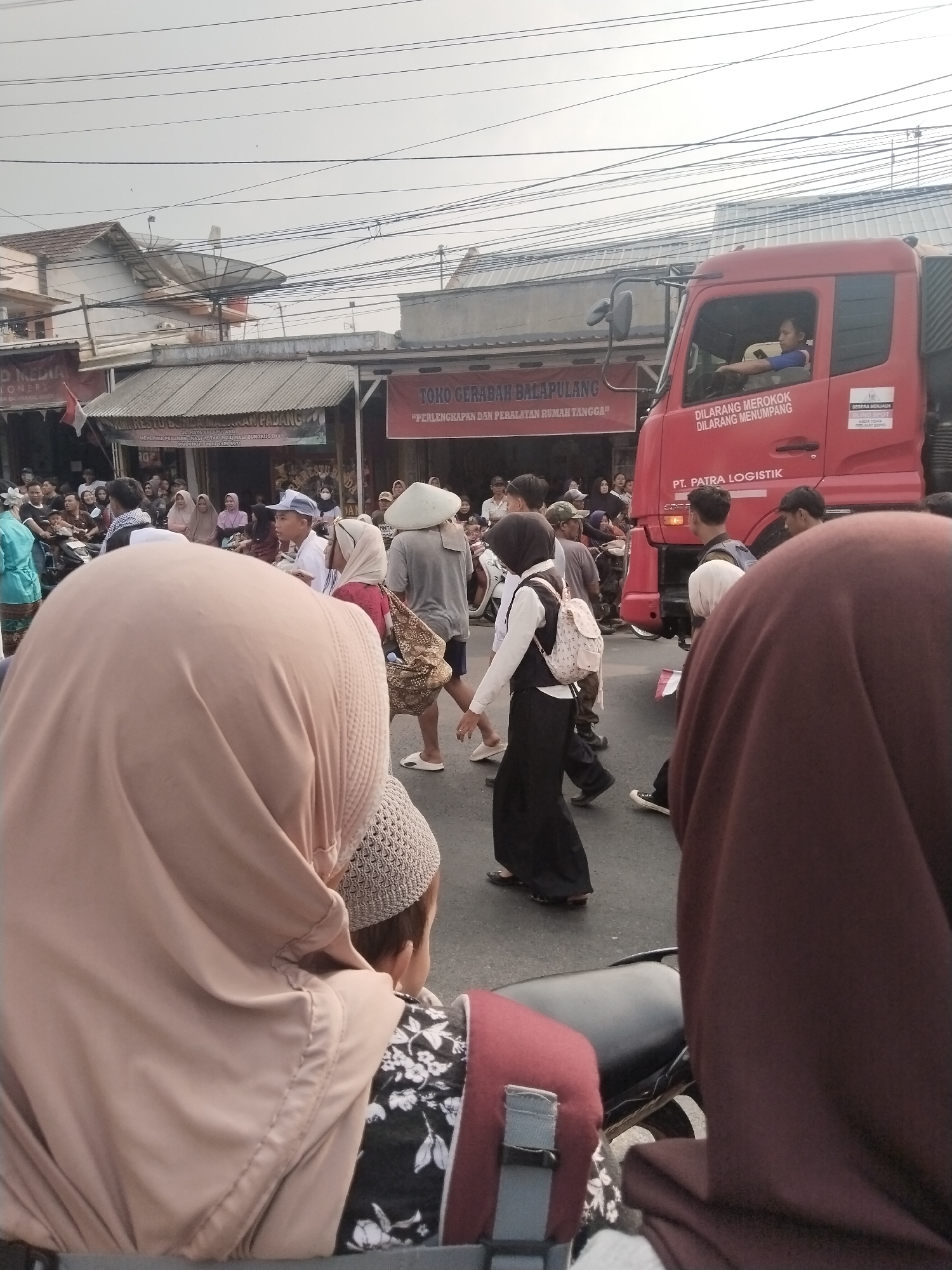 The image depicts a street scene in Balapulang Kulon, Indonesia, likely during a procession or event. A bright red truck with "PT. PATRA LOGISTIK" written on its side dominates the right side of the frame. The truck has a driver visible through the window, wearing a blue shirt. Various individuals are walking in the street, some wearing traditional Islamic attire like hijabs and peci caps. One person in the foreground is wearing a light brown hijab and a patterned garment. To the left, shops with awnings are visible, one of which has a banner that reads "TOKO GERABAH BALAPULANG". The sky is overcast. The ground is asphalt, and there are several power lines overhead. The overall atmosphere suggests a community gathering or a market day.
