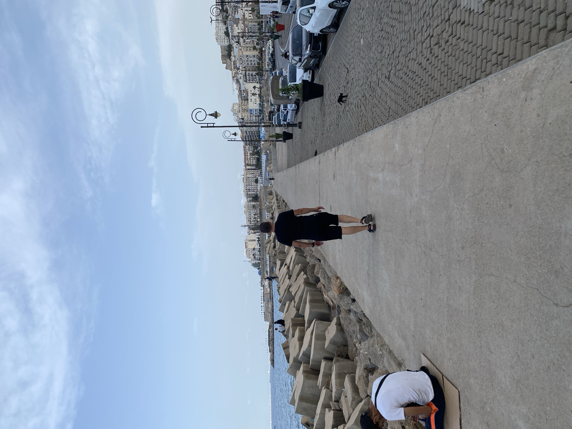 The image depicts a scenic waterfront promenade in Beau Fraisier, Algeria. The foreground shows a concrete walkway with a man in a black t-shirt and shorts walking away from the camera. To the left, large concrete blocks form a breakwater separating the walkway from the blue sea. A person in a white t-shirt and dark pants sits on a cardboard mat on the ground near the concrete blocks. In the background, a city with white buildings stretches along a hillside, with lampposts and parked cars lining the road adjacent to the walkway. The sky is a pale blue with scattered clouds.