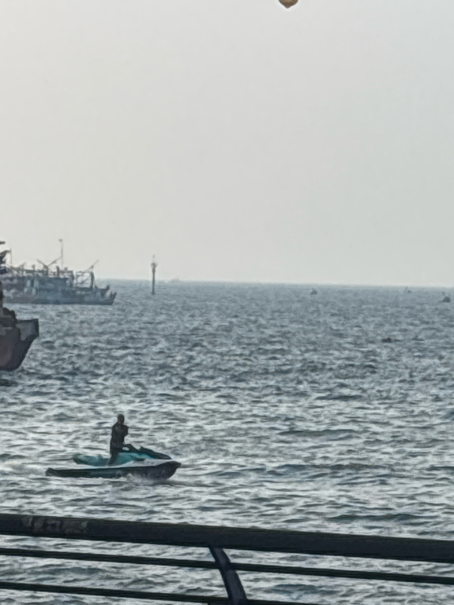 An overhead shot shows a person riding a jet ski in the water. The jet ski is blue and has a dark-colored person on it. The person is wearing a dark-colored shirt and pants. The jet ski is moving towards the left of the image. In the background, there are several boats. One boat is a large ship with several decks and cranes. There is also a buoy in the water. The sky is overcast and the water is choppy. The scene appears to be set in a harbor or port.