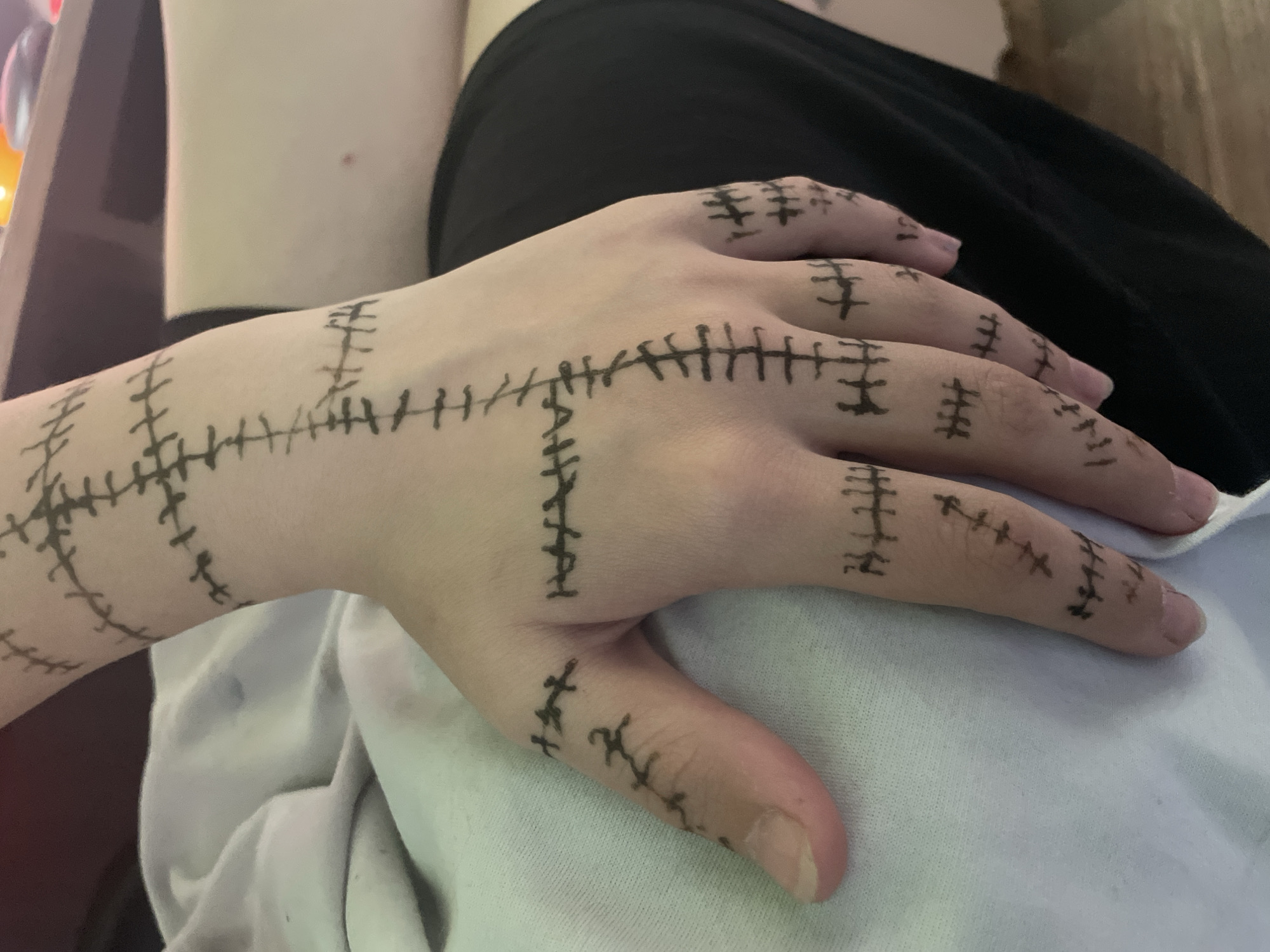 A person's right hand and a portion of their forearm are prominently displayed, covered in numerous black marker drawings designed to resemble stitches or sutures. These patterns consist of long lines intersected by many short, perpendicular dashes, creating the illusion of sewn-up skin. The markings extend across the back of the hand, along each finger, and up the visible part of the forearm. The individual's skin is fair, and their nails are natural and unpolished.
The hand rests on a soft, light-colored fabric, likely white, which could be a piece of clothing or a blanket. Behind the hand, to the left, a dark-colored garment, possibly black, is visible. The background is softly blurred, showing hints of an indoor setting, including another light-toned surface to the upper left. The lighting is diffused and even, suggesting an indoor environment rather than direct outdoor light. There is no discernible text present in the image. The activity appears to be the showcasing of temporary body art, possibly for a creative project, costume, or personal expression. No specific emotions are conveyed by the hand alone, but the markings evoke themes often associated with horror, patching, or unique aesthetic choices.