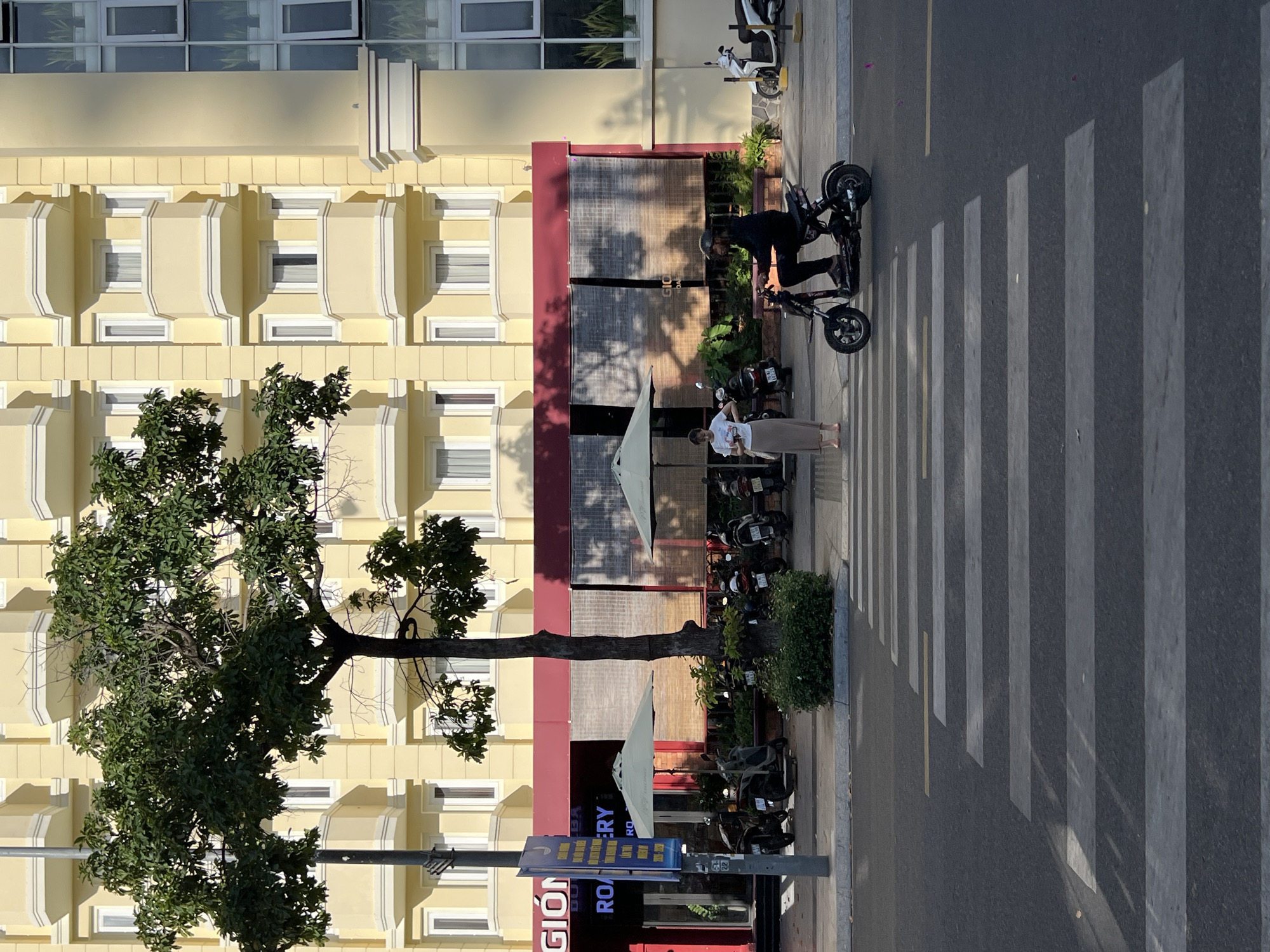 The image shows a street scene in Nha Trang Ward, Vietnam, with a yellow building dominating the background. In the foreground, a pedestrian crossing is visible on the asphalt road. A woman stands on the left side of the crossing, looking down at her phone. To her right, a person on an electric scooter is crossing the street, also from left to right. Several parked motorcycles are lined up along the sidewalk, behind the woman and the scooter. Trees and umbrellas are also present on the sidewalk in front of a building with red accents. The building has multiple windows and balconies.