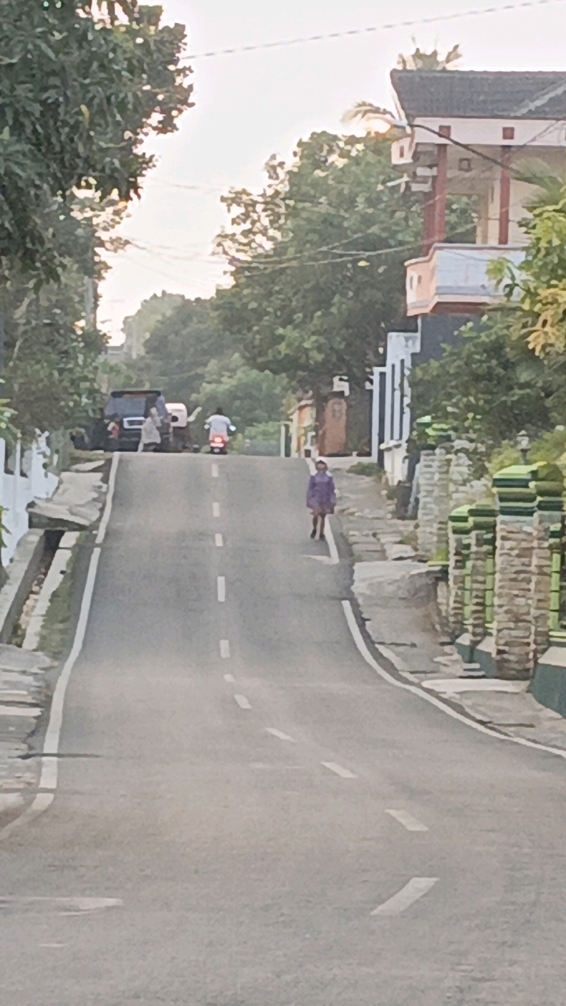 A slightly blurry, vertical, eye-level outdoor shot captures a residential street in Salatiga, Indonesia, during the day. The road, with a white dashed line dividing lanes, curves uphill. On the right side of the road, a person wearing a purple jacket and dark pants walks towards the camera. Beyond them, a wall constructed of stone blocks with green decorative elements lines the road. Further up the street, on the left, a dark SUV is parked, with two people standing near it. A motorcycle rider in white apparel is approaching the SUV from the top of the hill. Trees with dense foliage are visible on both sides of the street and in the background. Two-story houses with balconies and tiled roofs are situated behind the stone wall on the right. The sky is bright and appears to be overcast or hazy.