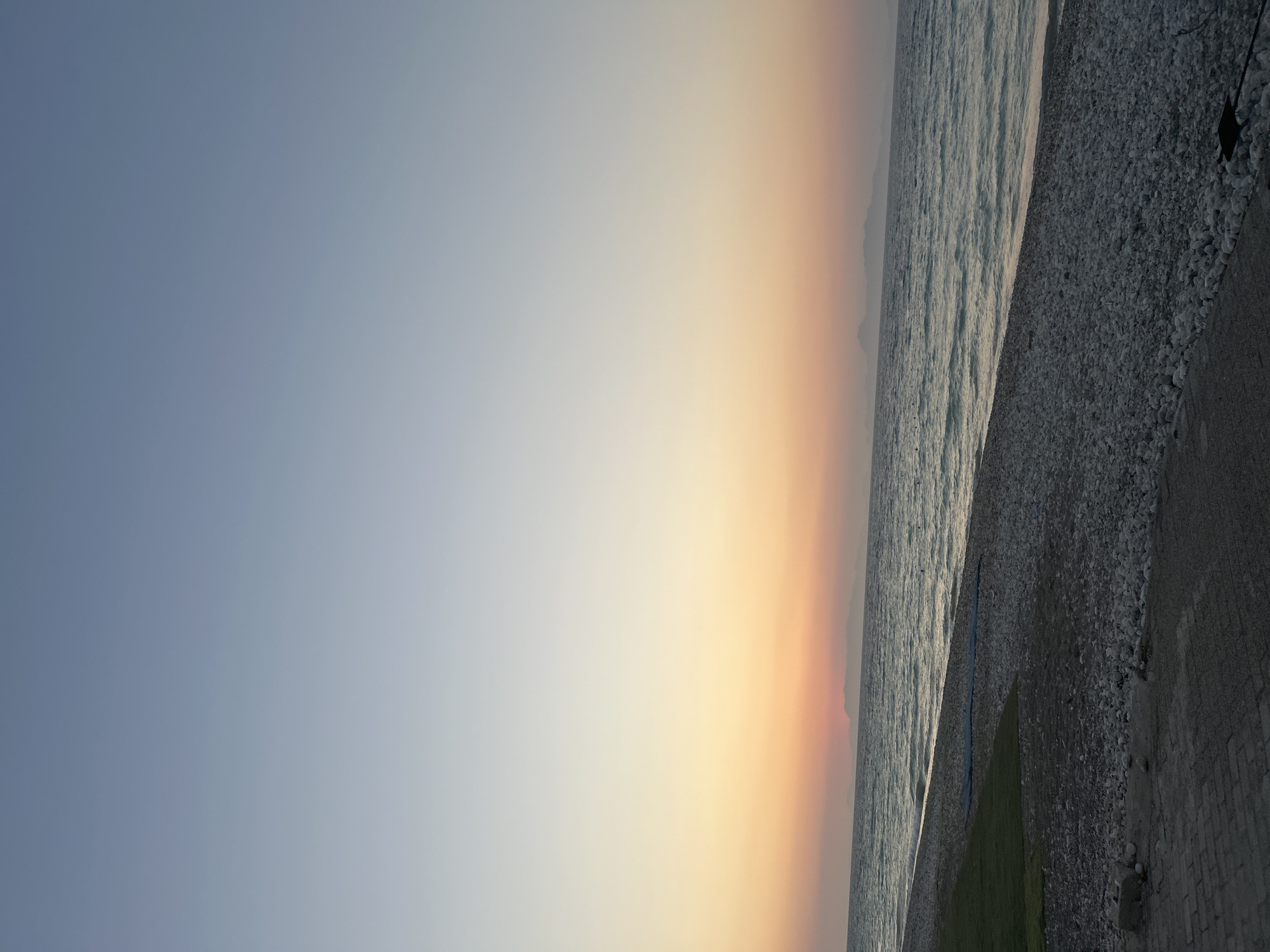 This is a landscape image of a serene beach at dusk. The image captures the gradient of a sunset sky, transitioning from a soft blue to warm hues of orange and yellow, indicating the time of day is either sunrise or sunset. On the right side of the frame, the calm, wavy sea meets a pebbled beach. The pebbles are varied in size and color, creating a textured surface. Further inland, there appears to be a patch of green grass and a paved pathway or promenade, suggesting an accessible coastline. In the far distance, the faint outline of hills or mountains can be seen against the horizon. The overall mood of the image is tranquil and peaceful, typical of a quiet beach scene during twilight. There is no visible text in the image.