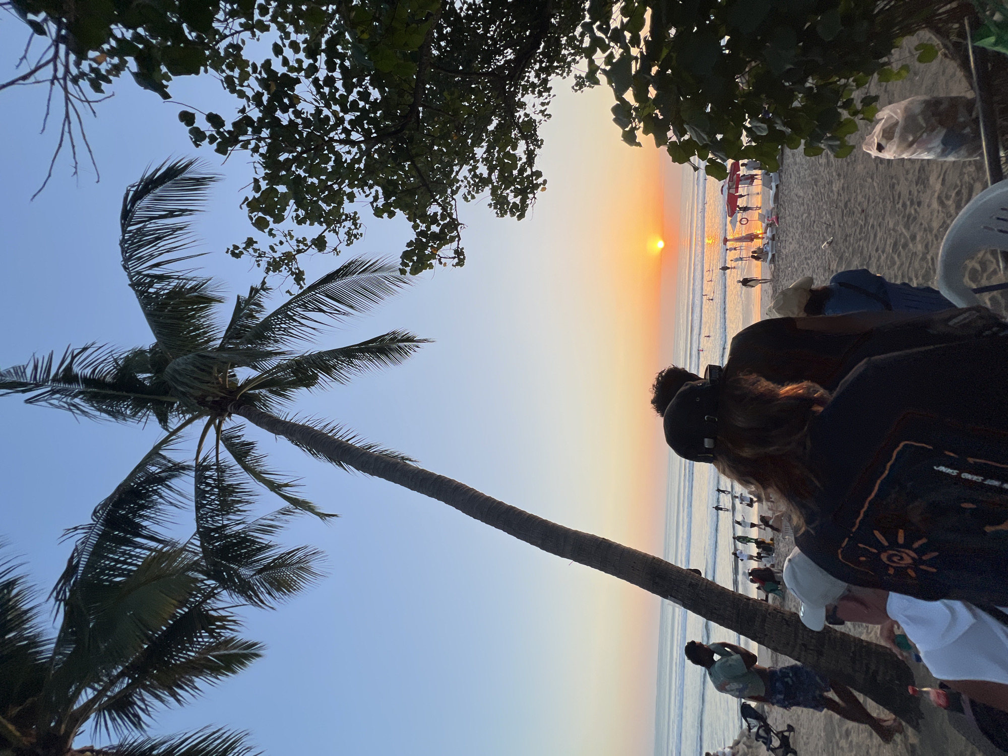 A group of people are gathered on the beach in Kuta, Indonesia, observing the sunset over the ocean. The sun is a bright orange orb low on the horizon, casting a warm glow across the water and sand. Palm trees frame the scene, with their fronds silhouetted against the clear, pale blue sky. In the foreground, several individuals are seated and standing, their backs mostly to the camera, suggesting they are engrossed in the view. Further down the beach, a few people are walking near the water's edge, and some are in the ocean, possibly swimming or surfing. Beach chairs and umbrellas are scattered on the sand, indicating a popular spot for relaxation. A trash bag is visible on the right side of the frame. The overall atmosphere is serene and peaceful, capturing a quintessential beach sunset experience.