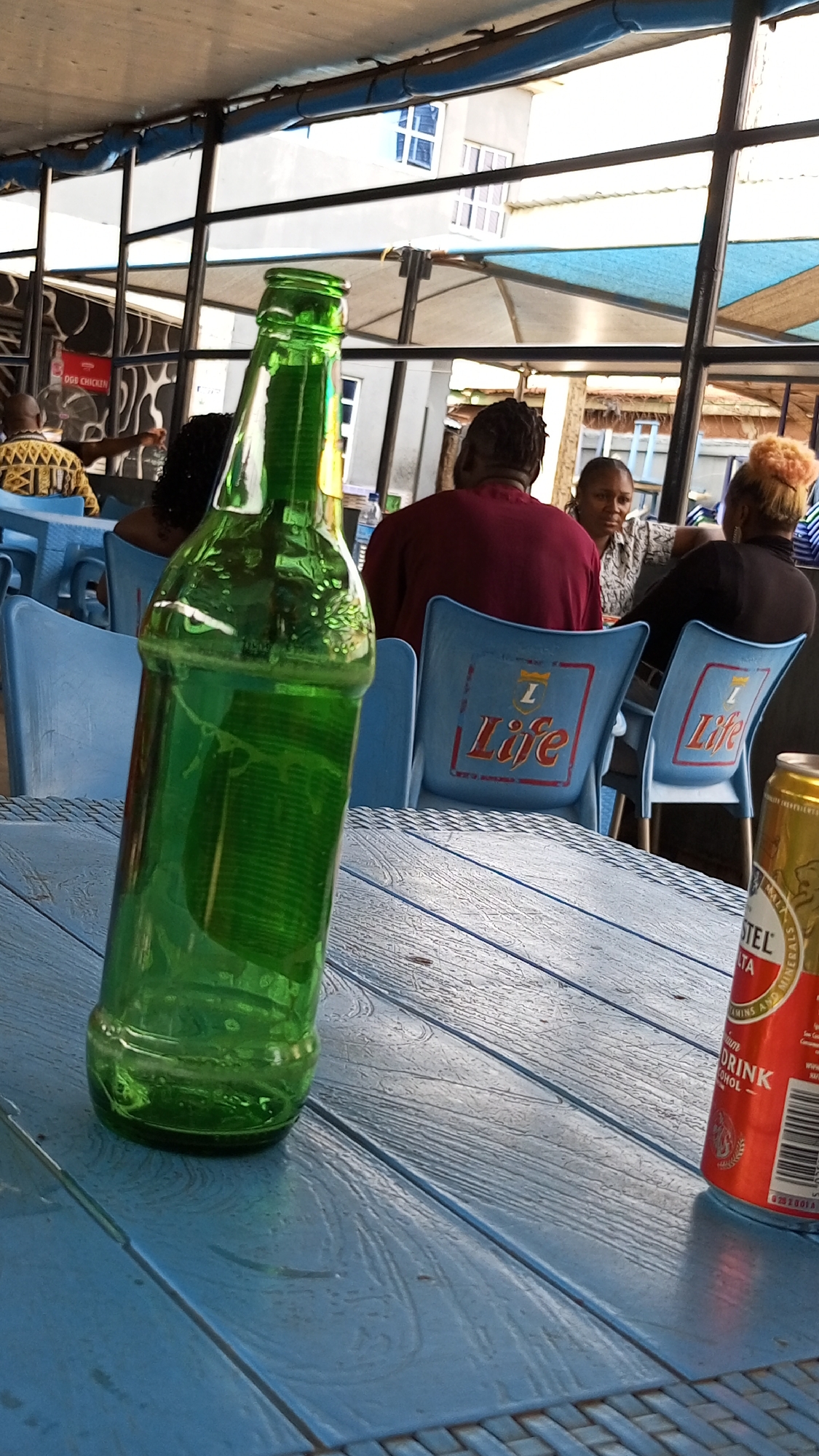 The image depicts an outdoor dining area, likely in Sabon Tasha, Nigeria. The scene is dominated by a light blue table, on which sits a nearly empty green glass bottle and a can of Amstel Malta. Several people are seated at tables in the background, seemingly engaged in conversation. The tables and chairs appear to be made of plastic, with some chairs bearing the word "Life" on their backs. The setting appears to be a semi-enclosed space with a view of buildings and the sky. The lighting suggests it is daytime. "OGB Chicken" is visible on a sign in the background.