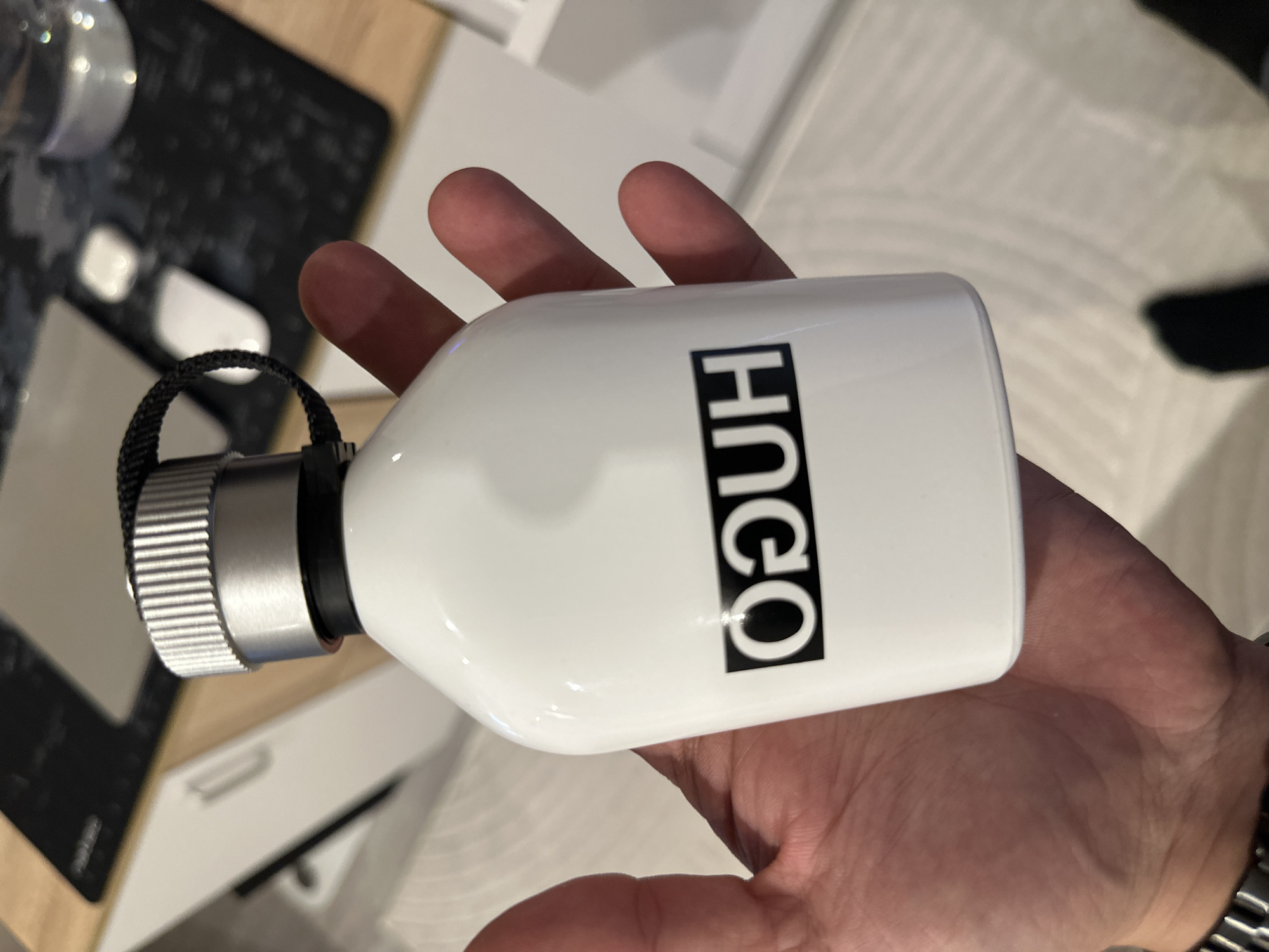 A person's hand holds a white water bottle with the word "HUGO" printed vertically in black letters. The bottle has a silver cap and a black loop attached. The background is a blurry mix of a desk with a black keyboard, a white surface, and a textured white floor. The photo was not taken in Lovech, Bulgaria.