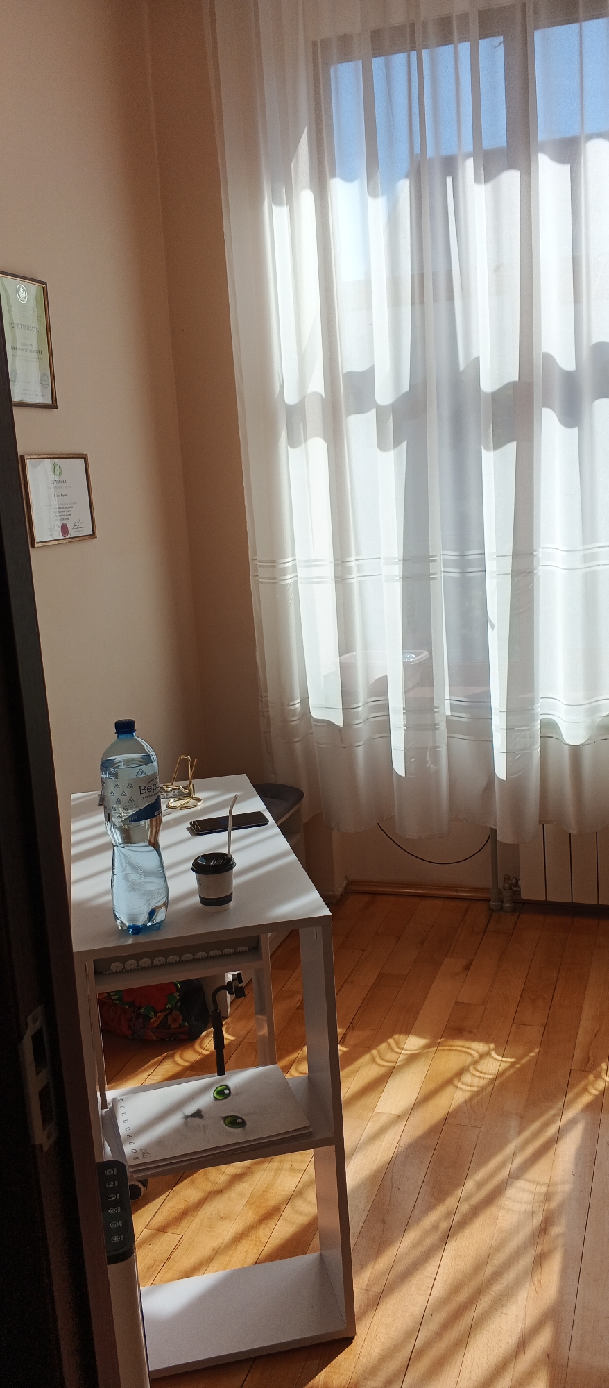 This is a medium shot, eye-level, indoor photograph taken in Mukachevo, Ukraine. The scene depicts a corner of a room with natural light streaming in through a large window covered by sheer white curtains. The sunlight casts distinct shadows across the light-colored wooden floor and the white desk in the foreground.
On the desk, there is a large plastic bottle of water, a black and white disposable coffee cup with a straw, and a smartphone. A gold-colored binder clip is also visible. A portion of a white shelving unit is present to the left of the desk. On the top shelf of this unit, a notebook with a drawing of a cat's face with green eyes is resting. The bottom shelf is empty.
On the wall to the left of the desk, two framed certificates are hanging. The upper certificate is partially visible and has text including "CERTIFICATE". The lower certificate is more clearly visible and also appears to be a formal document with text and a seal.
The environment appears to be a study or office area. The overall mood is calm and quiet, emphasized by the natural light and the arrangement of objects.