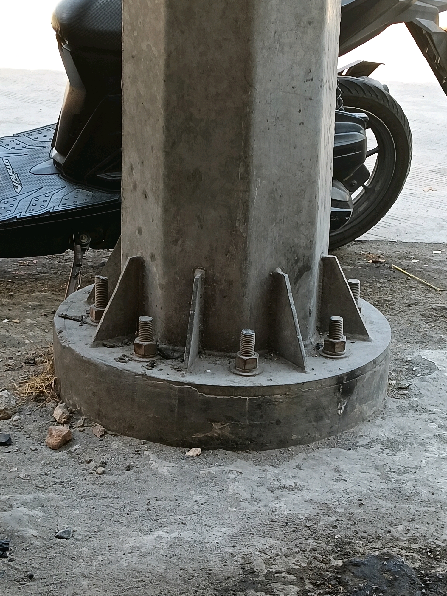 A close-up, eye-level shot focuses on the base of a concrete utility pole, anchored to the ground by large bolts. A portion of a black scooter is visible to the left of the pole, its textured footrest and seat suggesting it is parked. To the right, the rear wheel and engine area of a motorcycle are partially visible. The ground surrounding the pole is composed of rough, grey concrete and gravel, with scattered small stones and dry grass. The lighting suggests it is daytime, with soft shadows indicating diffused sunlight. The scene is set outdoors, likely on a street or pavement in Kedungtuban, Indonesia, given the context. No individuals are present in the frame.
