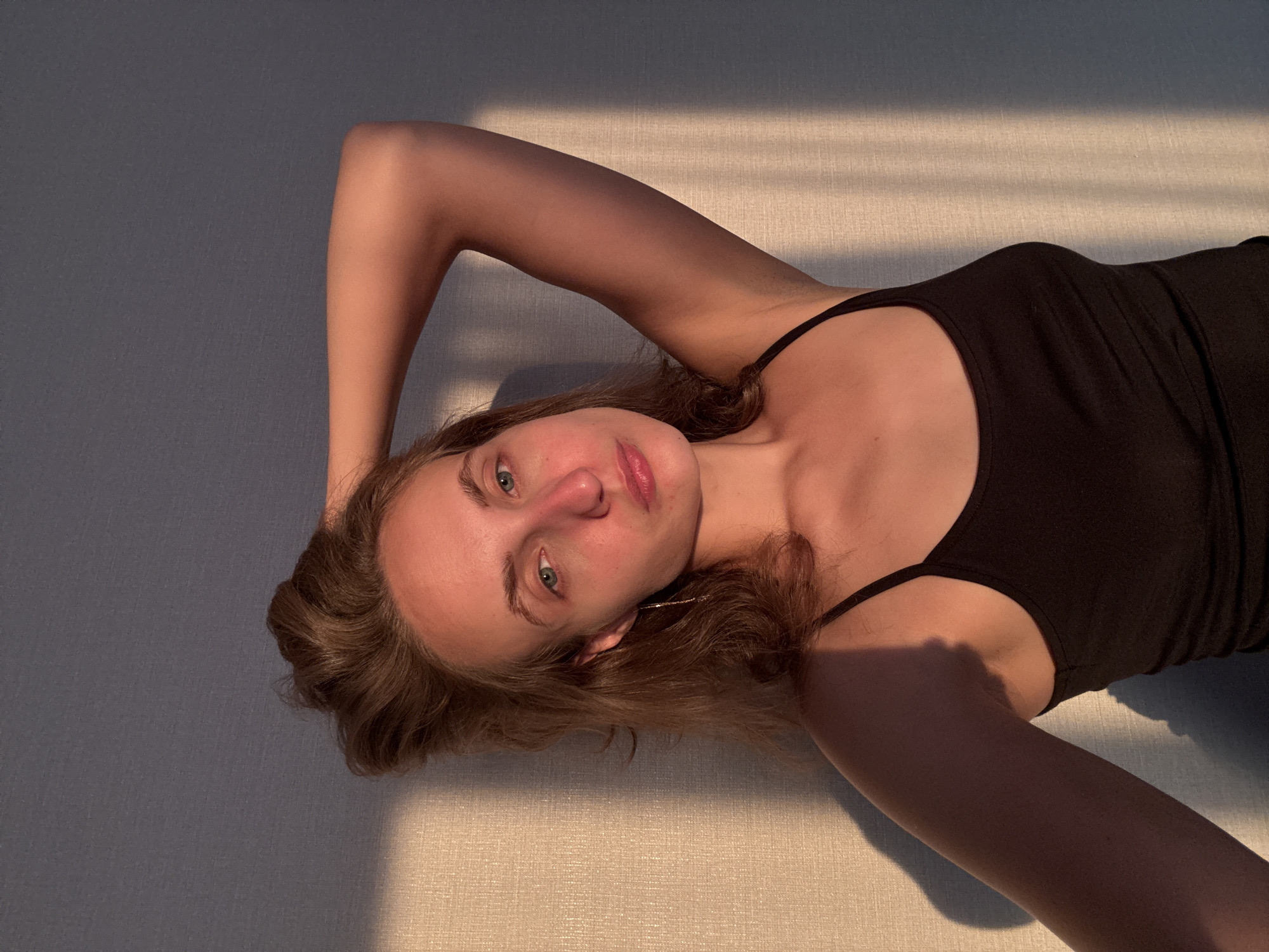 A young Caucasian woman is shown in a close-up, frontal portrait, bathed in the warm, angled light of sunset. She has light brown hair, pulled back slightly from her face, and blue eyes. Her expression is neutral, with her head tilted slightly and her right arm raised behind her head. She is wearing a black tank top. The background is a textured wall, with strong vertical shadows cast by an unseen object. The lighting suggests an indoor setting, possibly an apartment in the city of Podshivalovo, Russia.