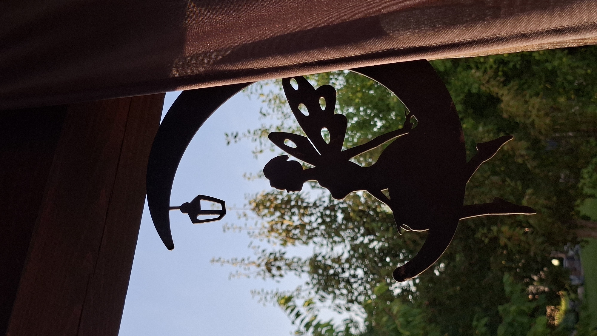 A silhouetted metal ornament of a fairy sitting on a crescent moon with a lantern hanging above it is positioned in front of a blurred background of green trees against a clear blue sky. The ornament is dark brown and appears to be made of metal. The fairy has delicate wings with cut-out shapes and is seated with her legs dangling over the moon's curve. A rough-textured, dark brown fabric curtain hangs on the right side of the image, partially obscuring the scene. The ornament is mounted on a dark wooden beam that runs along the top of the frame. The image is taken outdoors, likely on a sunny day, with the golden hour sunlight illuminating the edges of the ornament. The location is specified as Myadzyel, Belarus.