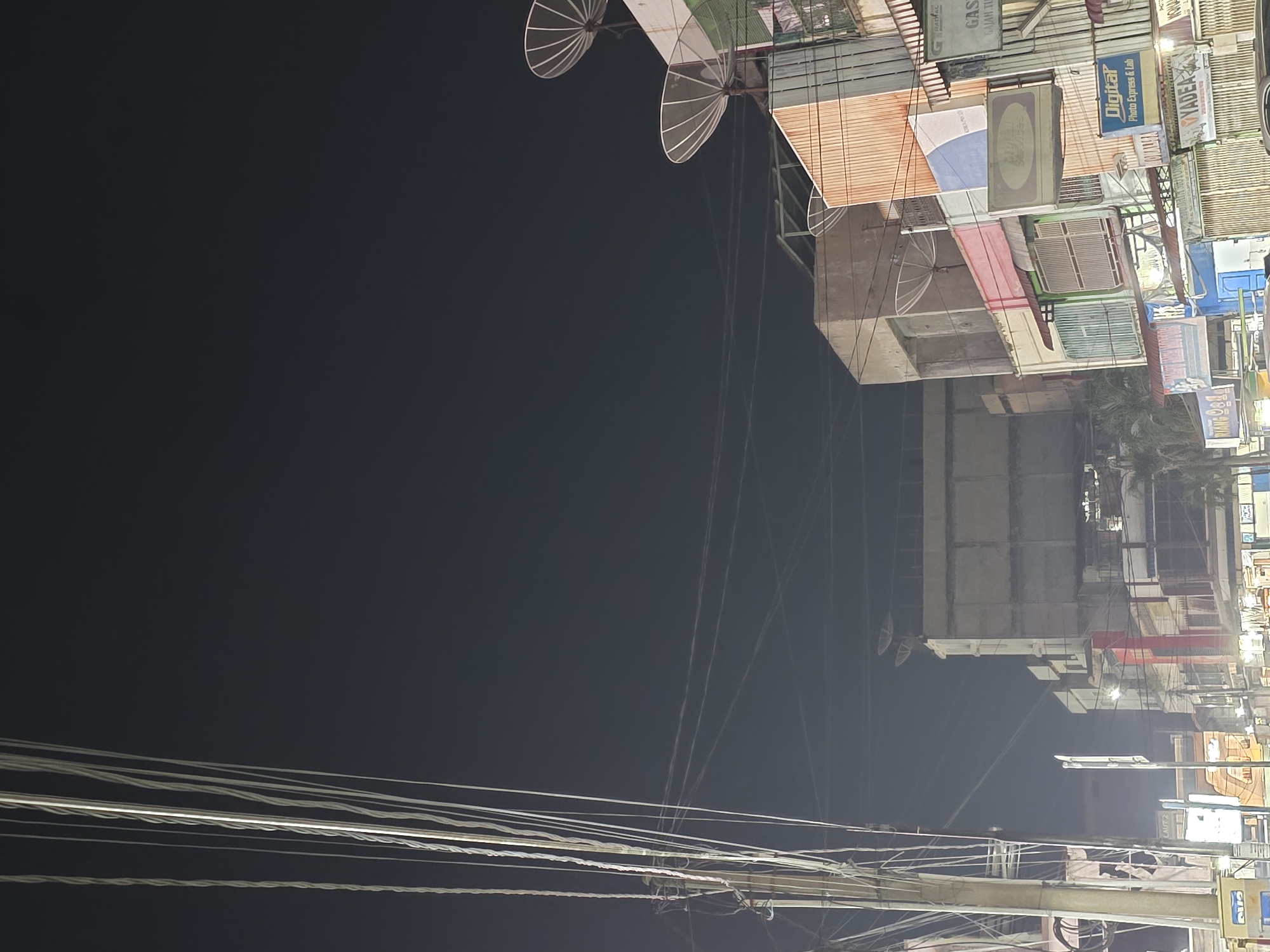 The image shows a street scene at night in the city of Bireuen, Indonesia. The foreground is dominated by a utility pole with numerous wires. In the background, several multi-story buildings line the street, some of which appear to be shops or businesses. One building has several satellite dishes mounted on its roof. Signs in Indonesian are visible on some of the buildings, including "Digital Photo Express & Lab" and "YADEA". The overall atmosphere is that of a typical urban street at night. There are no people visible in the image.