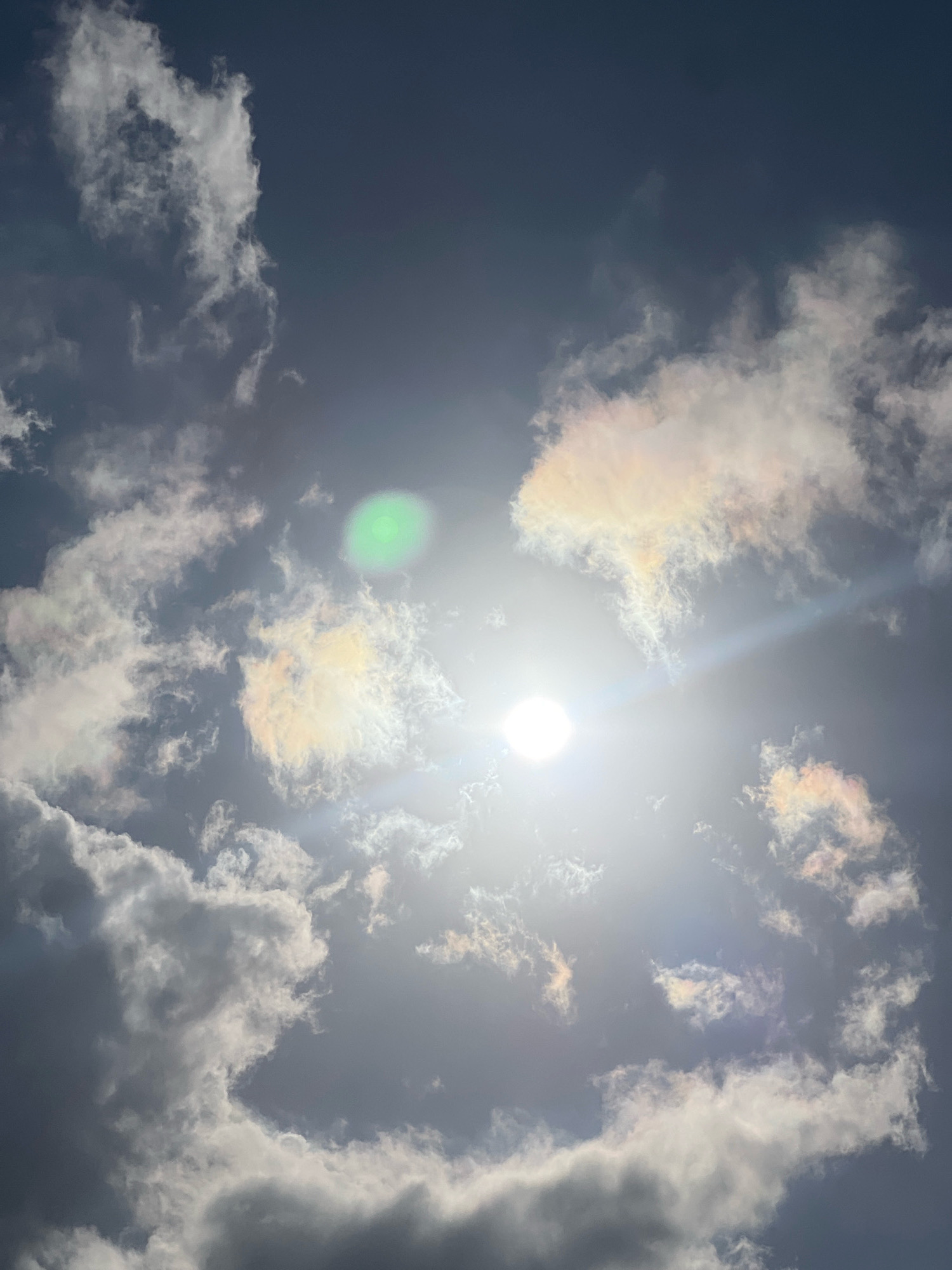The image shows a bright sun in a partly cloudy sky. The sun is located in the lower right quadrant of the image, emitting a strong glare that obscures details. There are several clouds scattered throughout the frame, some appearing bright white and illuminated by the sun, while others are darker and more shadowed. Some of the clouds exhibit iridescence, displaying subtle rainbow-like colors. A faint green lens flare is visible in the upper left portion of the image. The sky appears to be a deep blue or grey, suggesting either early morning, late afternoon, or an overcast condition with the sun breaking through. There are no people, buildings, or any identifiable landmarks to suggest a specific location within Denpasar, Indonesia. The image is solely focused on the sky and clouds.