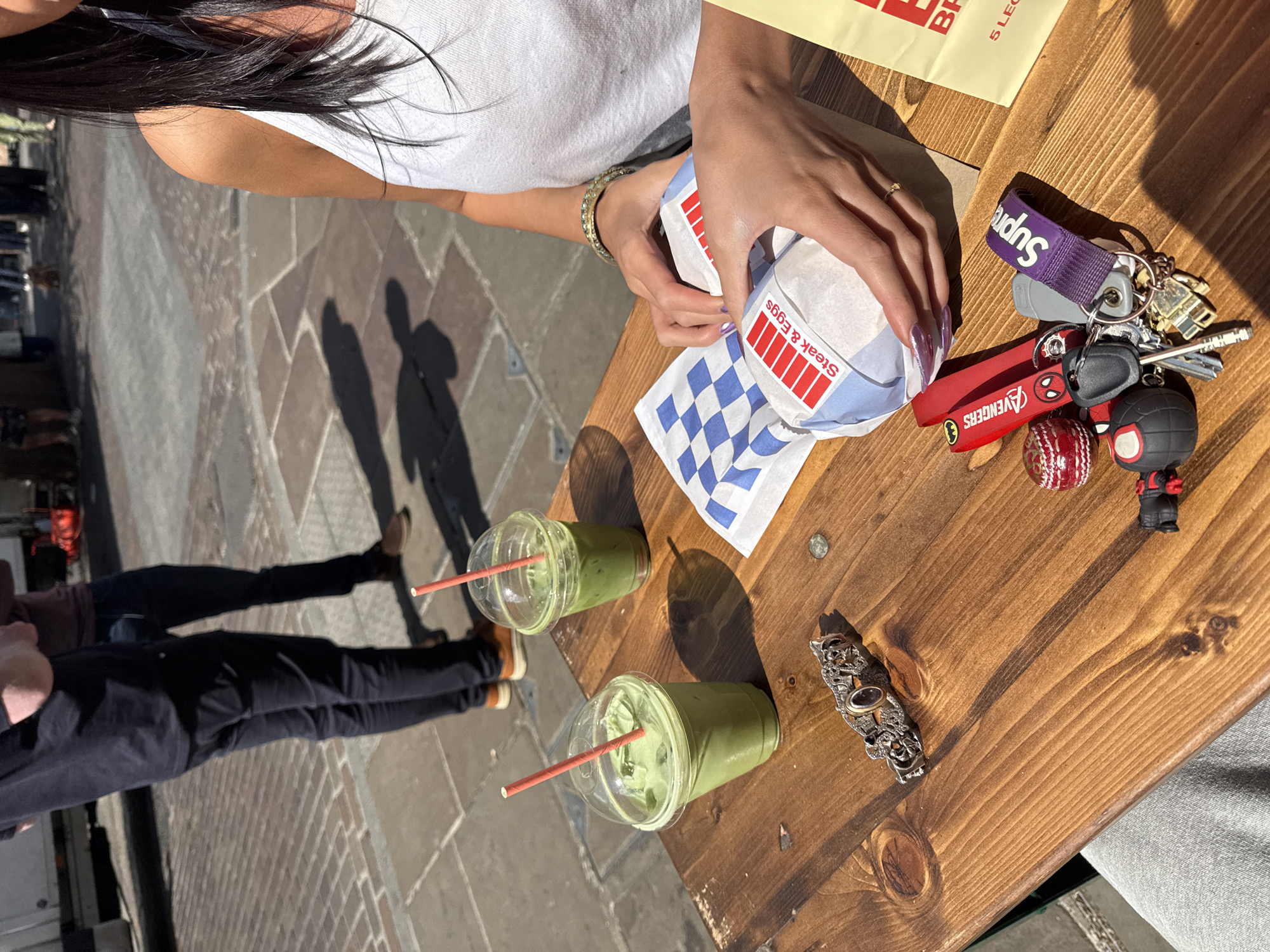 The image captures an outdoor dining scene on a sunny day. A person, partially visible with long dark hair and wearing a white top, is seated at a light brown wooden table, holding a food item wrapped in paper. Their hands are engaged with the packaging, which features red and white stripes and the text "Steak & Eggs." Another part of the food packaging shows a blue and white checkered pattern.
On the table, two clear plastic cups containing green beverages, likely matcha lattes, are visible, each with a red straw. To the right of the person's hand, a collection of keys is laid out. These keys are attached to a purple lanyard displaying the word "Supreme," a red keychain with "Avengers" text, a red cricket ball-shaped keychain, and a Spider-Man head keychain. An ornate silver-colored metal object, possibly a decorative buckle or bracelet, also rests on the table. A yellow paper, possibly a menu or receipt, with "5 LEG" visible, is also present.
In the background, two individuals are standing on a paved surface, characterized by large, square tiles or cobblestones. Their figures are largely seen as prominent, long shadows stretching across the ground, indicating bright sunshine from a particular angle. Their lower bodies, dressed in dark trousers and shoes, are also visible. The strong shadows on the ground and table confirm it is a clear, sunny day. The setting appears to be a casual outdoor eating area in an urban environment.