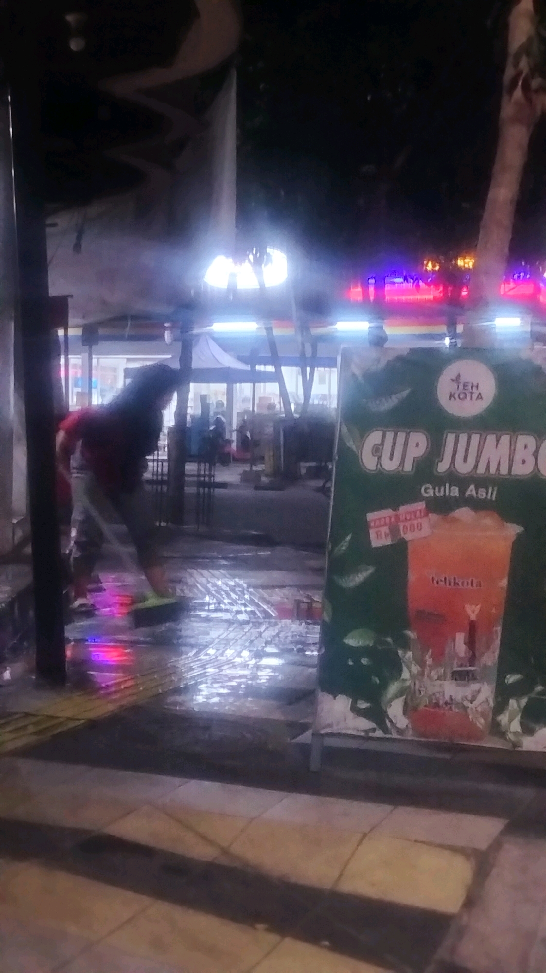 An individual is sweeping water on a wet, paved sidewalk at night. The person, wearing a dark top and light-colored pants, uses a broom to move a puddle of water to the right. In the background, there are buildings with bright lights, and a large green advertisement sign is visible on the right side of the image. The sign features text and an image of a drink. The overall scene appears to be an outdoor area in a city at night, likely near shops or food stalls. The location is specified as Bojonegoro, Indonesia.