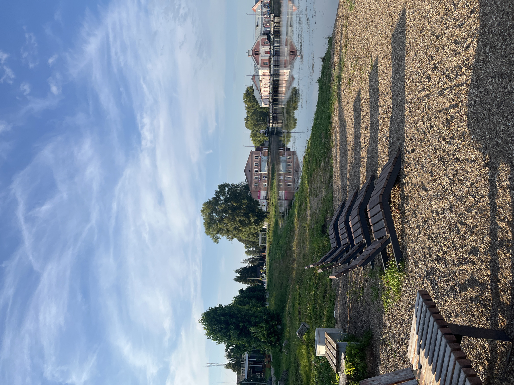 The image depicts a waterside scene in Ufa, Russia. In the foreground, several empty wooden chaise lounges are arranged on a gravelly surface, with one wooden bench partially visible on the left. Behind the chaise lounges, a grassy area slopes down towards a body of water. Across the water, several buildings are visible. One building on the left is a light brown, two-story structure with windows. Further to the right is a larger, pink and white building with a long arcade and arched windows, connected to another pink and white structure. These buildings are reflected in the calm water, which also shows some scattered debris on its surface. The sky above is a clear blue with scattered wispy white clouds. Trees line the far bank of the water, and a tall communication tower is visible in the distance on the left. The overall impression is a quiet, outdoor public space, likely a park or waterfront promenade, in a city environment.