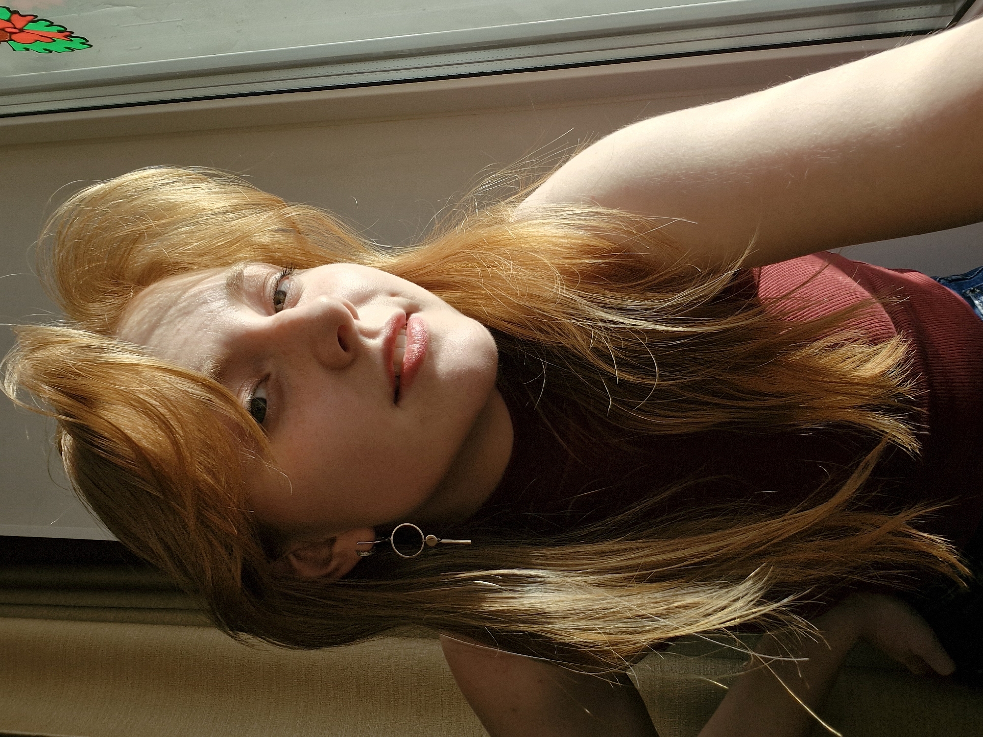 A young woman with long, flowing reddish-blonde hair is the primary subject of this indoor portrait, taken in Yekaterinburg, Russia. She is positioned near a window, likely lying down or reclining on a light brown textured surface, possibly a carpet or blanket. Her head is tilted, and she looks directly at the camera with a relaxed, gentle smile, her lips slightly parted.
She has light-colored eyes, possibly green or hazel, and a fair complexion. She is wearing a dark red or maroon ribbed top, and a portion of blue denim fabric, likely jeans, is visible in the lower right. A distinct silver earring with a black circular center and an elongated bar adorns her left ear.
The scene is brightly lit by direct sunlight streaming through the window, casting distinct shadows on her face and hair, indicating it is daytime. A decorative sticker, appearing as a green leaf with red elements, is visible on the window glass in the upper left corner. No text is visible in the image. The overall mood conveyed is warm, natural, and serene.