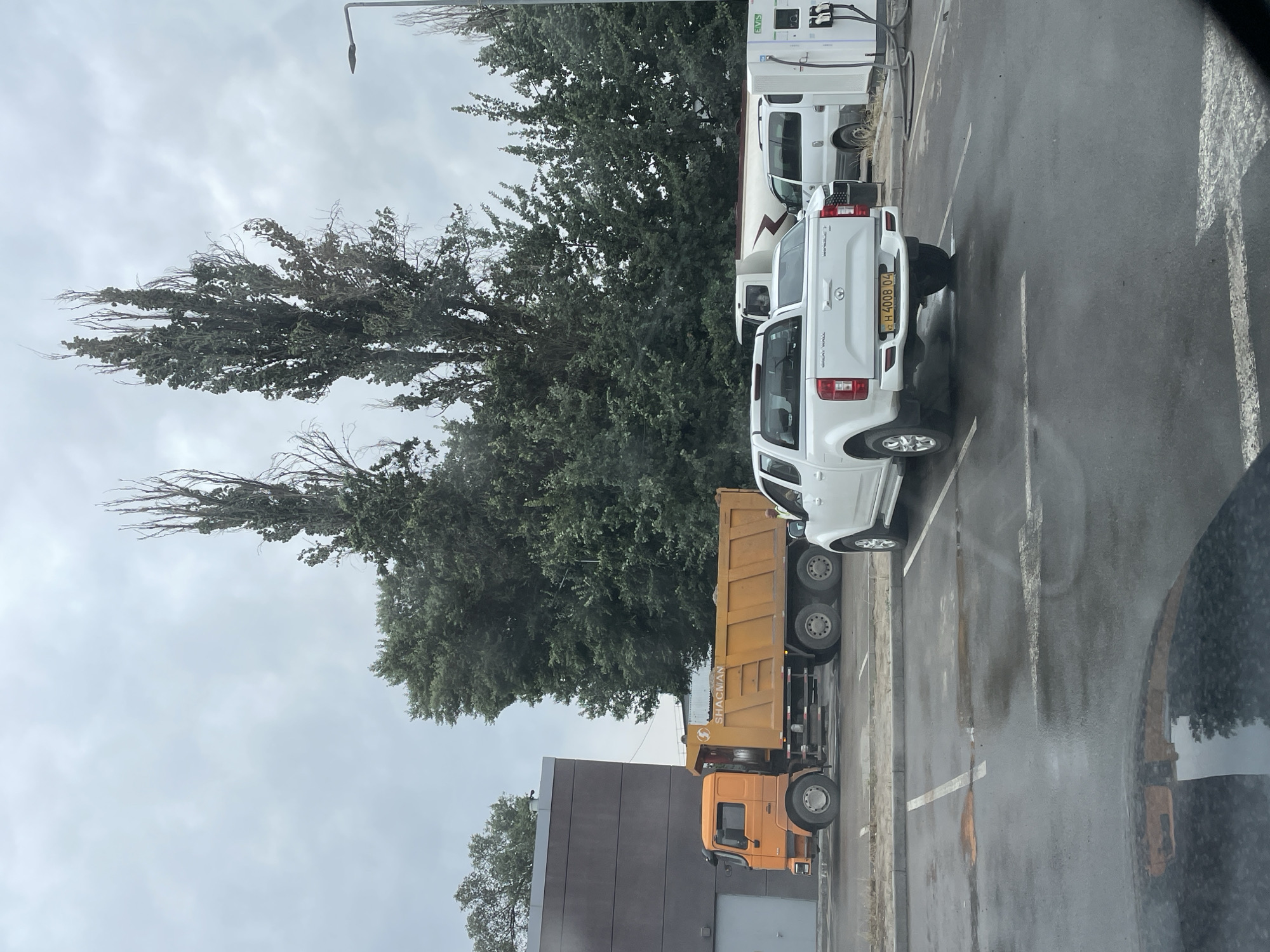 In Almaty, Kazakhstan, a dump truck is parked next to a building. A white pickup truck is parked behind the dump truck. Further back, there are two white vans and charging stations. The sky is overcast.