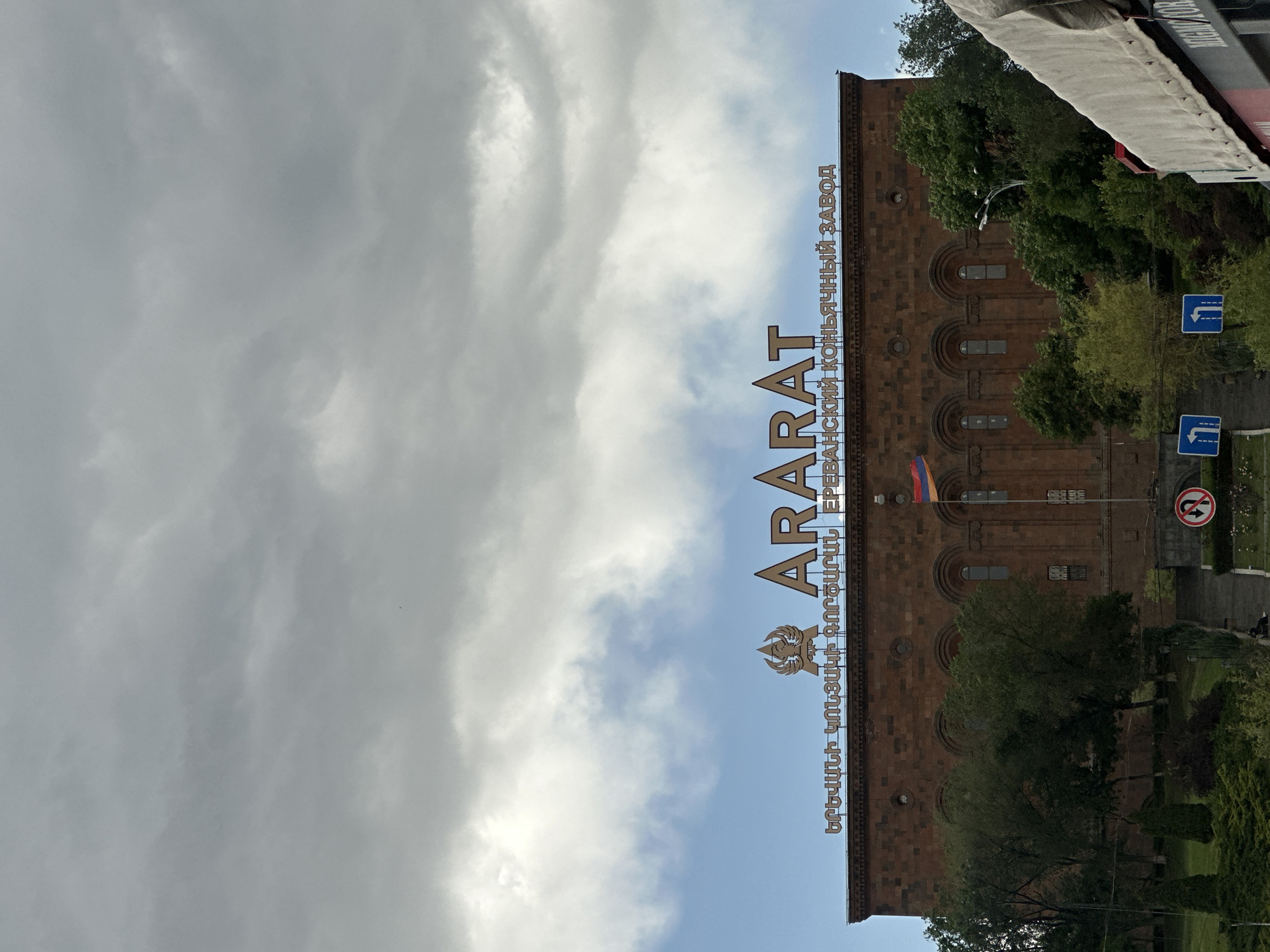 The image shows the Yerevan Ararat Brandy Factory in Yerevan, Armenia. The building is a multi-story brick structure, featuring arched windows and a flag. The words "ARARAT" and "ԵՐԵՎԱՆԻ ԿՈՆՅԱԿԻ ԳՈՐԾԱՐԱՆ" (Yerevani Konyaki Gortsaran, meaning Yerevan Brandy Factory) are written in a golden font on the building, as well as "ЕРЕВАНСКИЙ КОНЬЯЧНЫЙ ЗАВОД" in Russian (Yerevanskiy Konyachnyy Zavod, meaning Yerevan Brandy Factory). The image is taken from a low angle, with a partly cloudy sky in the background. Trees and street signs are visible.