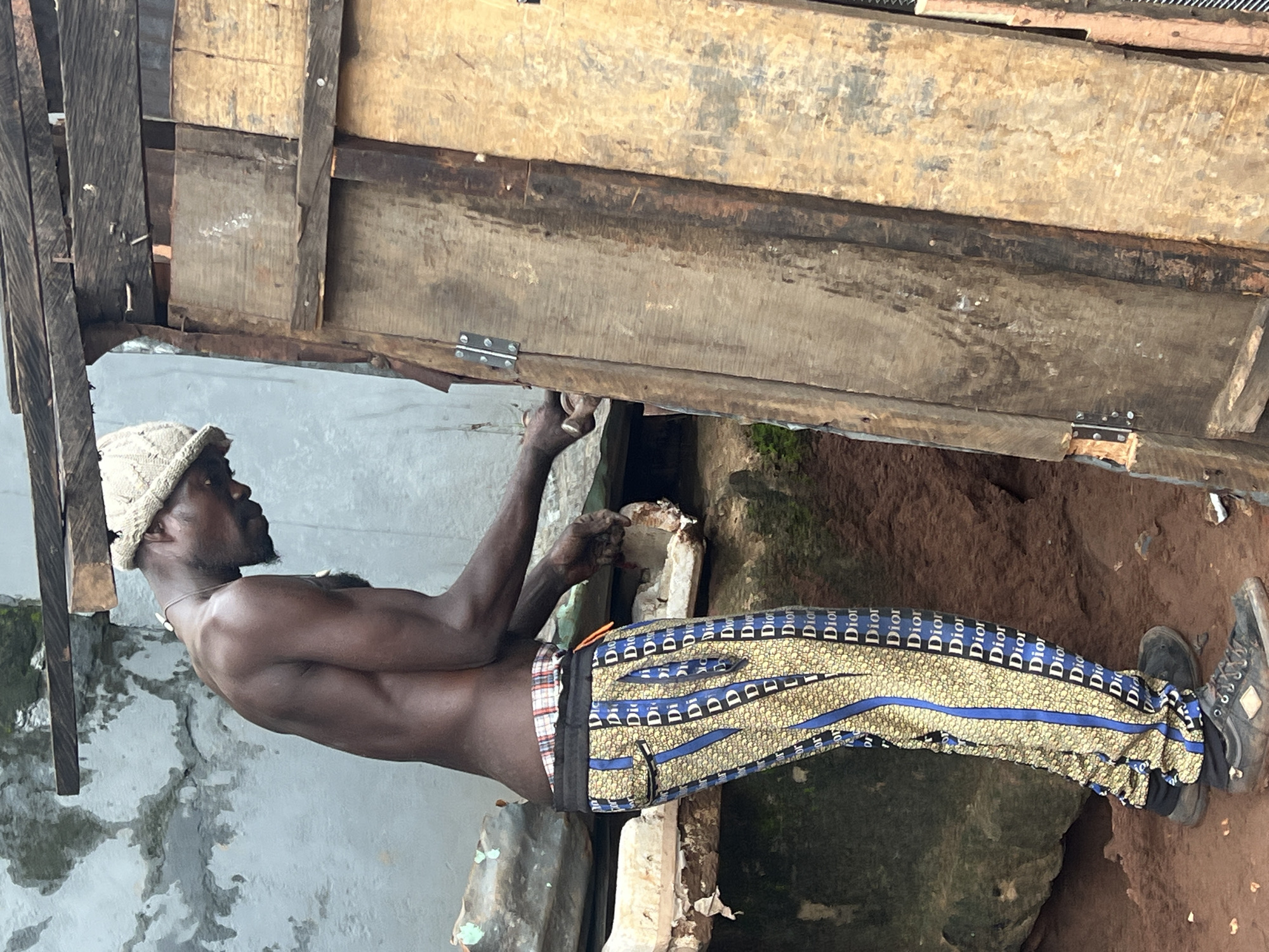 A dark-skinned man wearing a knit hat, a necklace, and patterned pants with the word "Dior" repeated on them is working on a wooden structure. He is shirtless, exposing his muscular back and arms. The man is positioned in what appears to be an outdoor, possibly construction or repair, setting in Nkpor, Nigeria. The ground is covered in reddish-brown soil. To his right, several rough, weathered wooden planks are visible, some forming a vertical structure. A metal hinge is attached to the wooden structure. In the background, a grey, textured wall suggests a building or dwelling. The overall scene depicts a moment of manual labor in a rustic environment.